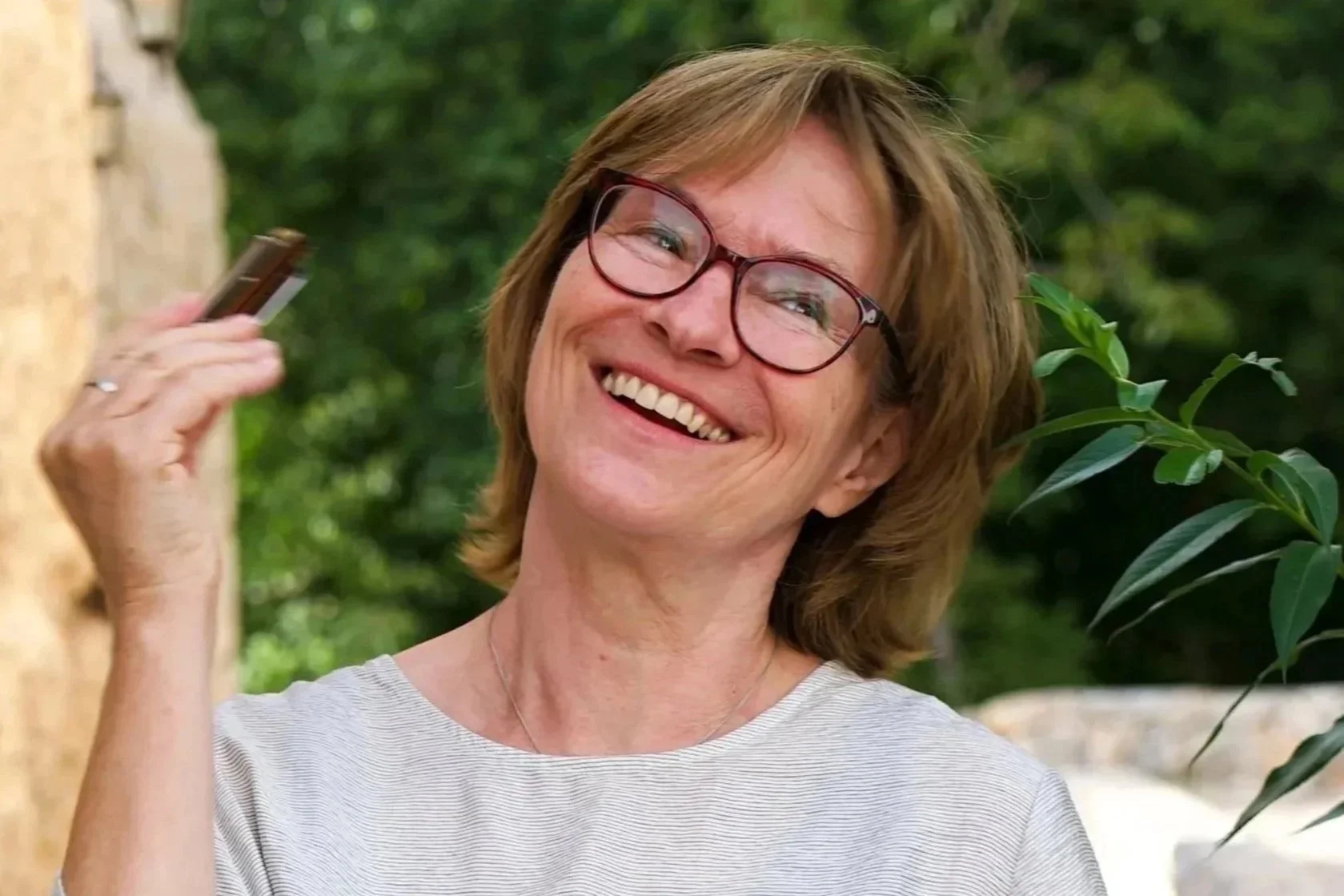 Lächelnde Frau mit Brille und kurzen braunen Haaren, die ein Handy in der Hand hält, draußen in einer grünen Umgebung.