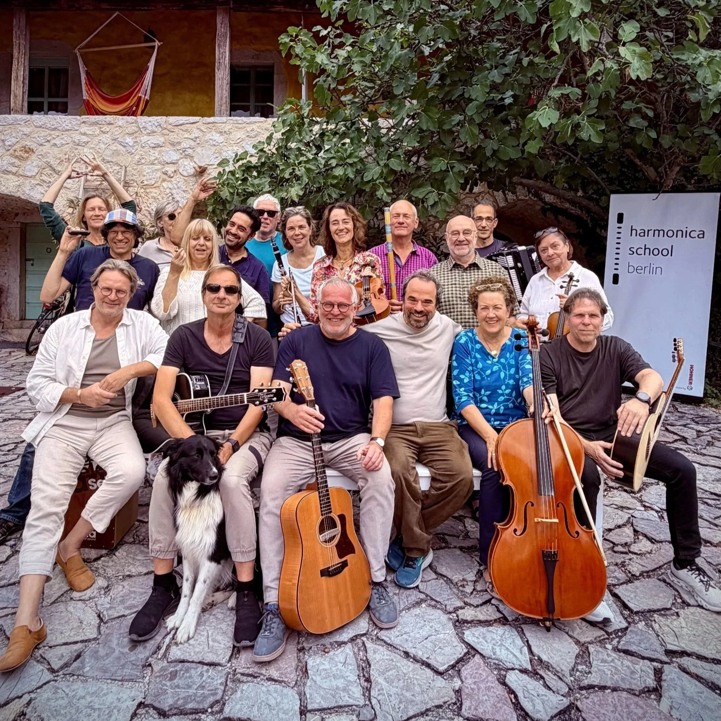 Gruppe von Musikern mit Instrumenten vor einem Baum und einem Gebäude, daneben ein Schild mit der Aufschrift 'harmonica school berlin'.