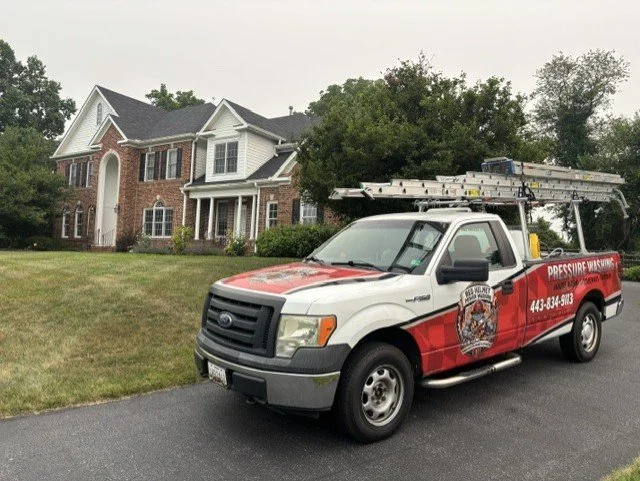 A white and red pressure washing service truck parked on a driveway in front of a large brick house with a well-maintained lawn and trees.
