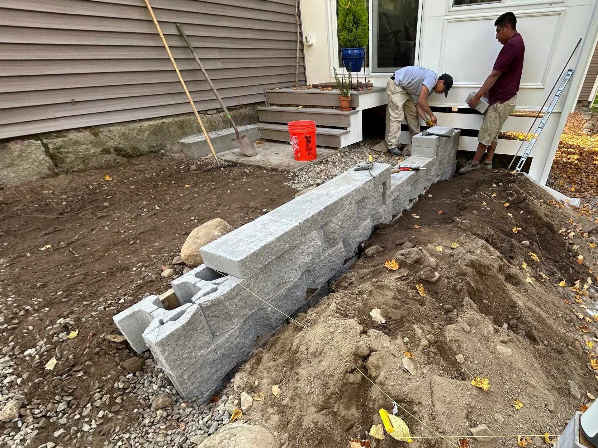 Two construction workers building a stone wall outside a house. One worker is bending over placing a stone, the other is holding a trowel. Construction tools and materials are scattered around.