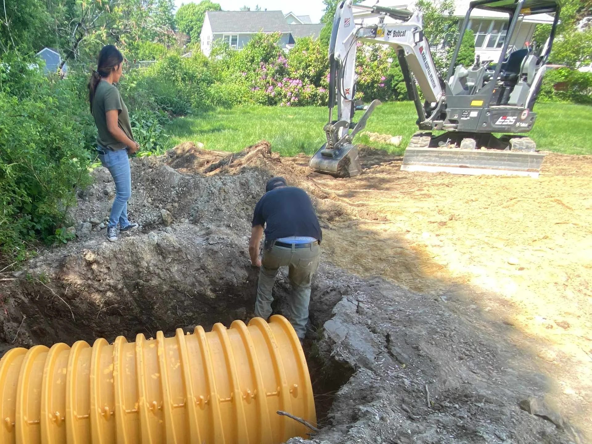 Two men and a woman working on a construction site outdoors, with a small excavator and a yellow drainage pipe, next to a grassy yard with trees and houses in the background.