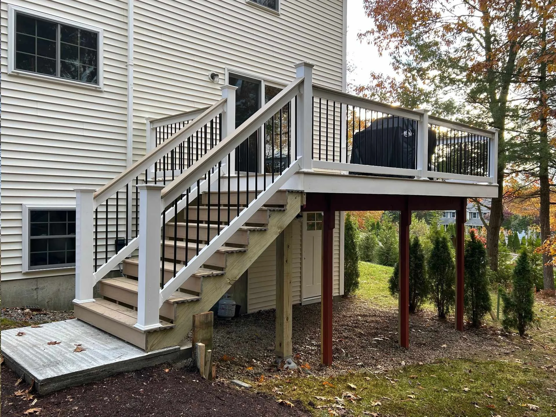 A newly built wooden deck with black railing on the back of a house, with stairs leading down to the yard. The house has beige siding and white-framed windows. There is a black grill on the deck. The yard has trees with fall foliage, and the ground i