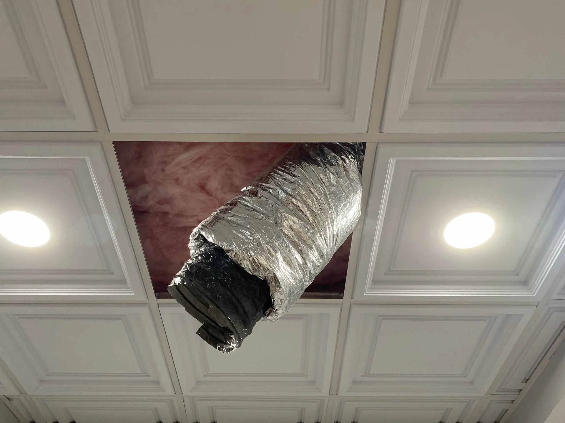 Ceiling with white decorative panels and two round recessed lights, with a metal HVAC duct hanging from the ceiling.