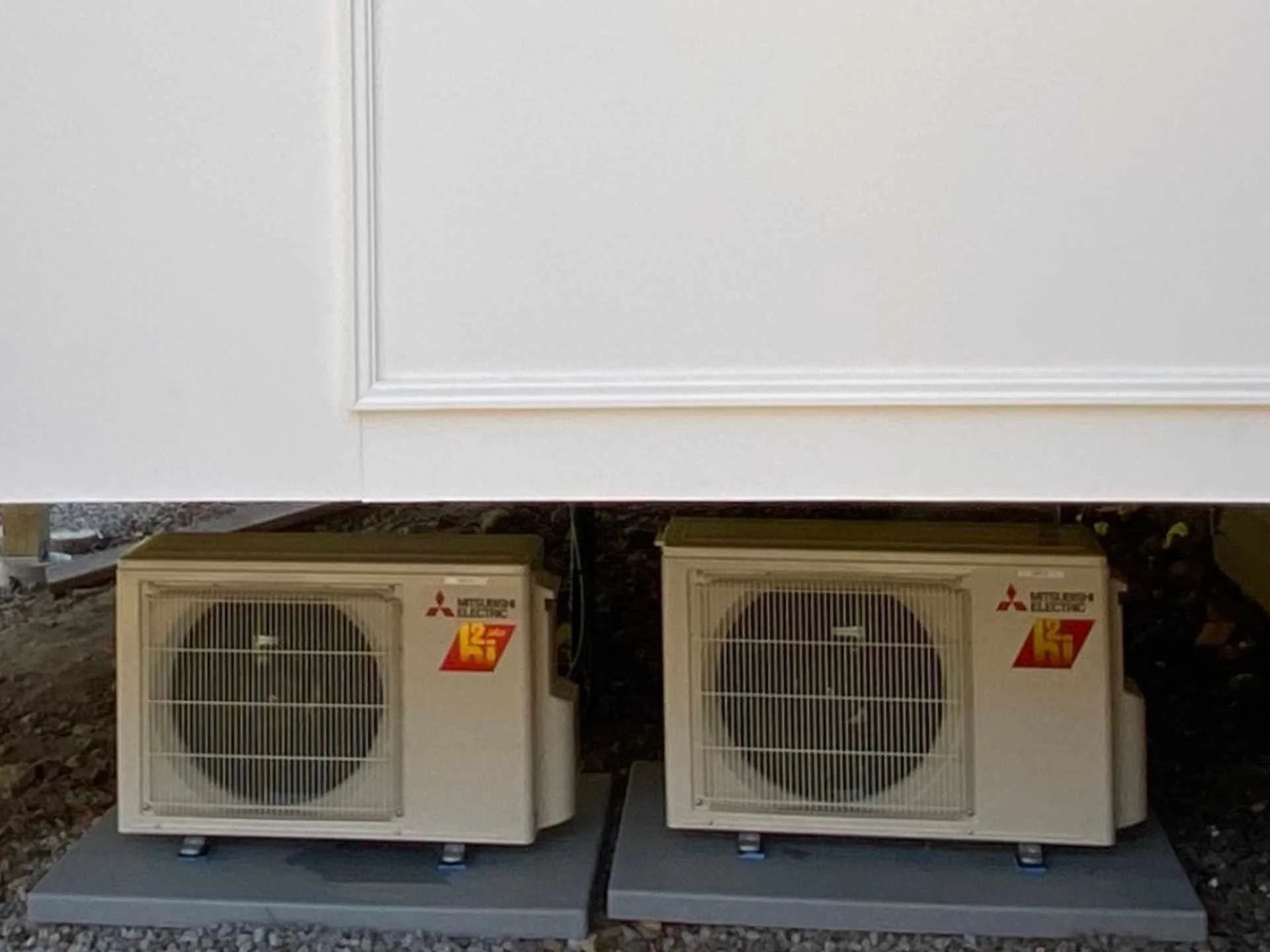 Two beige Mitsubishi air conditioning and heating units installed outdoors on a concrete platform beneath a white wall.