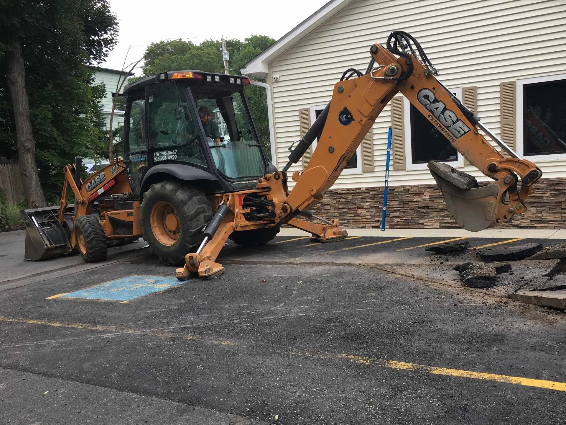 A backhoe loader excavator working on a parking lot near a beige Enterprise Rent-A-Car office with brown shutters. The parking lot has a designated disabled parking space with yellow lines, and some asphalt patches are being removed.