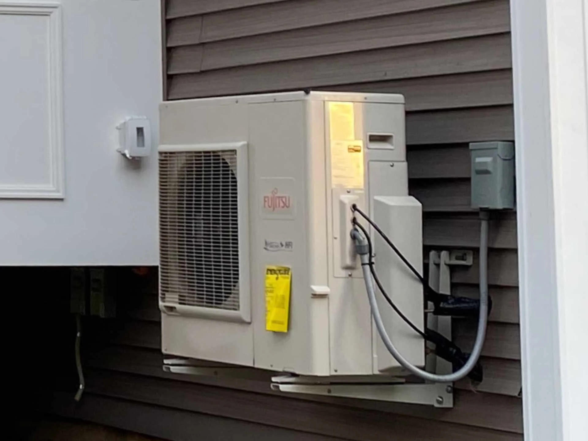 An air conditioning and heating unit mounted on an outdoor wall with a vinyl clapboard siding background.
