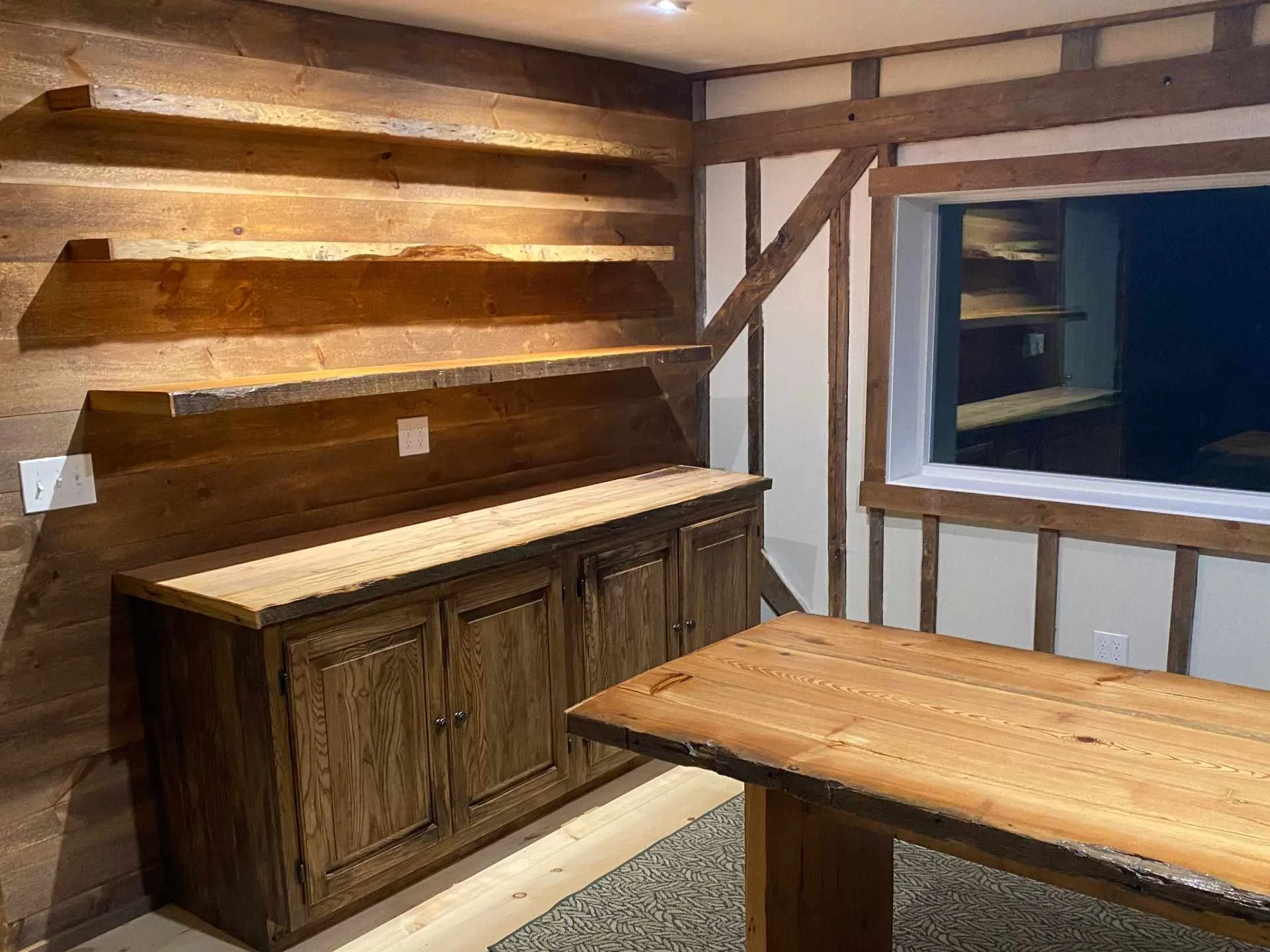 Interior of a room with rustic wooden wall paneling, a custom-built wooden cabinet, and a custom dining table made from reclaimed heart pine, with a window showing a reflection of the room. the exposed framing members of the rustic building add charm