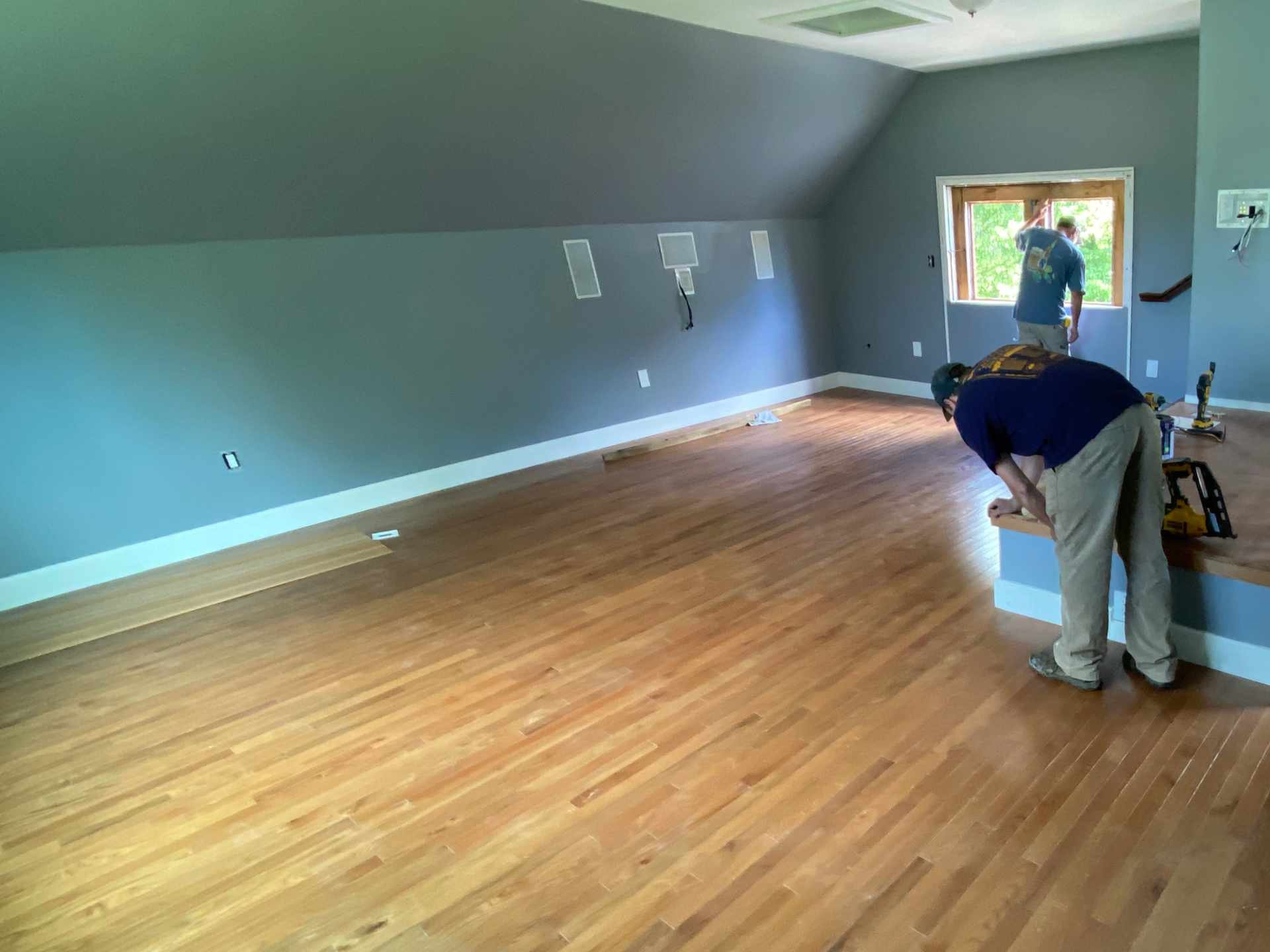 Workers installing hardwood flooring in a room with a sloped ceiling and a large window, with tools on the floor.