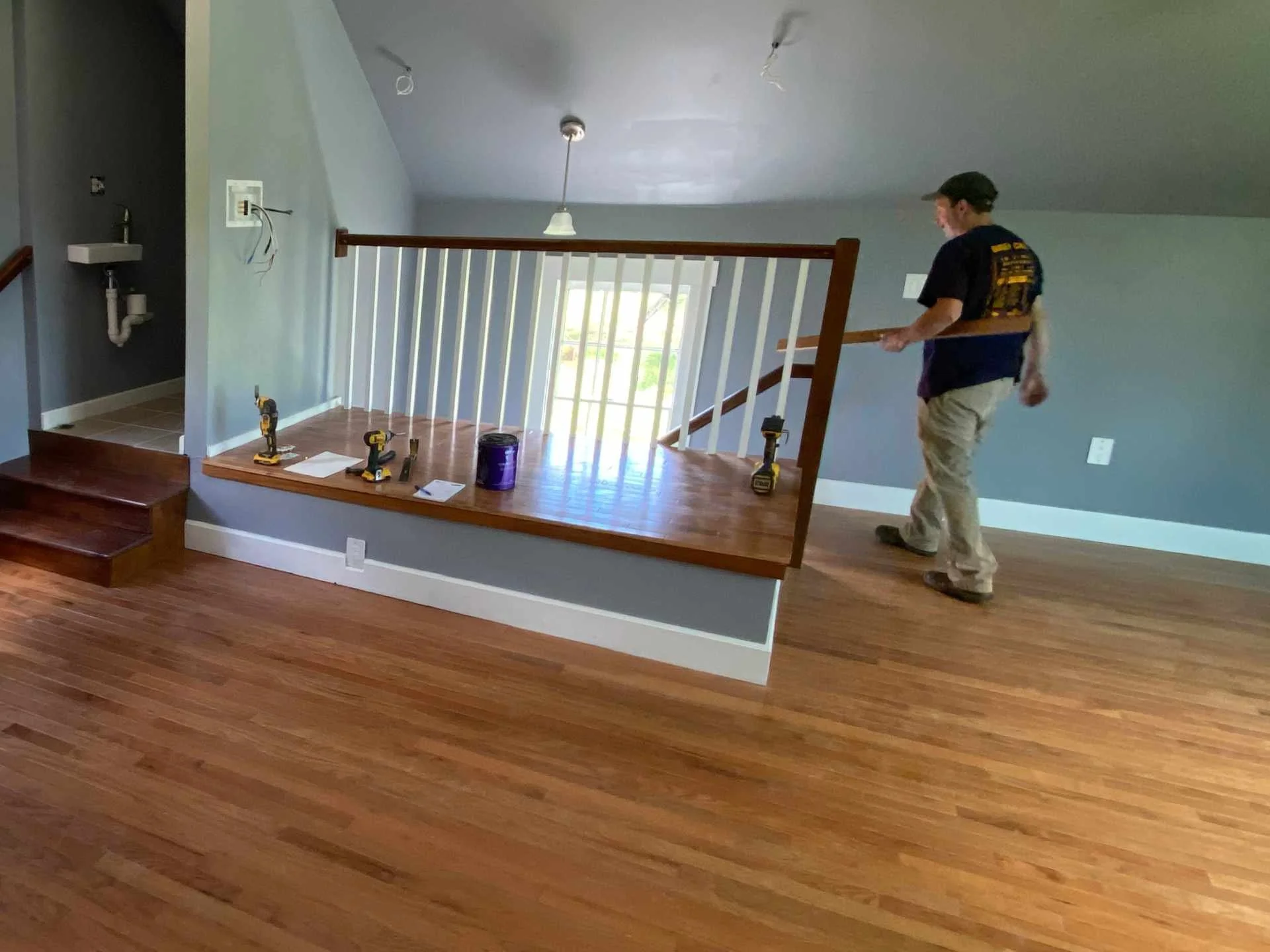 A person installing or repairing the stair railing of a staircase in a room with wooden floors and light blue walls, tools and supplies on the floor.