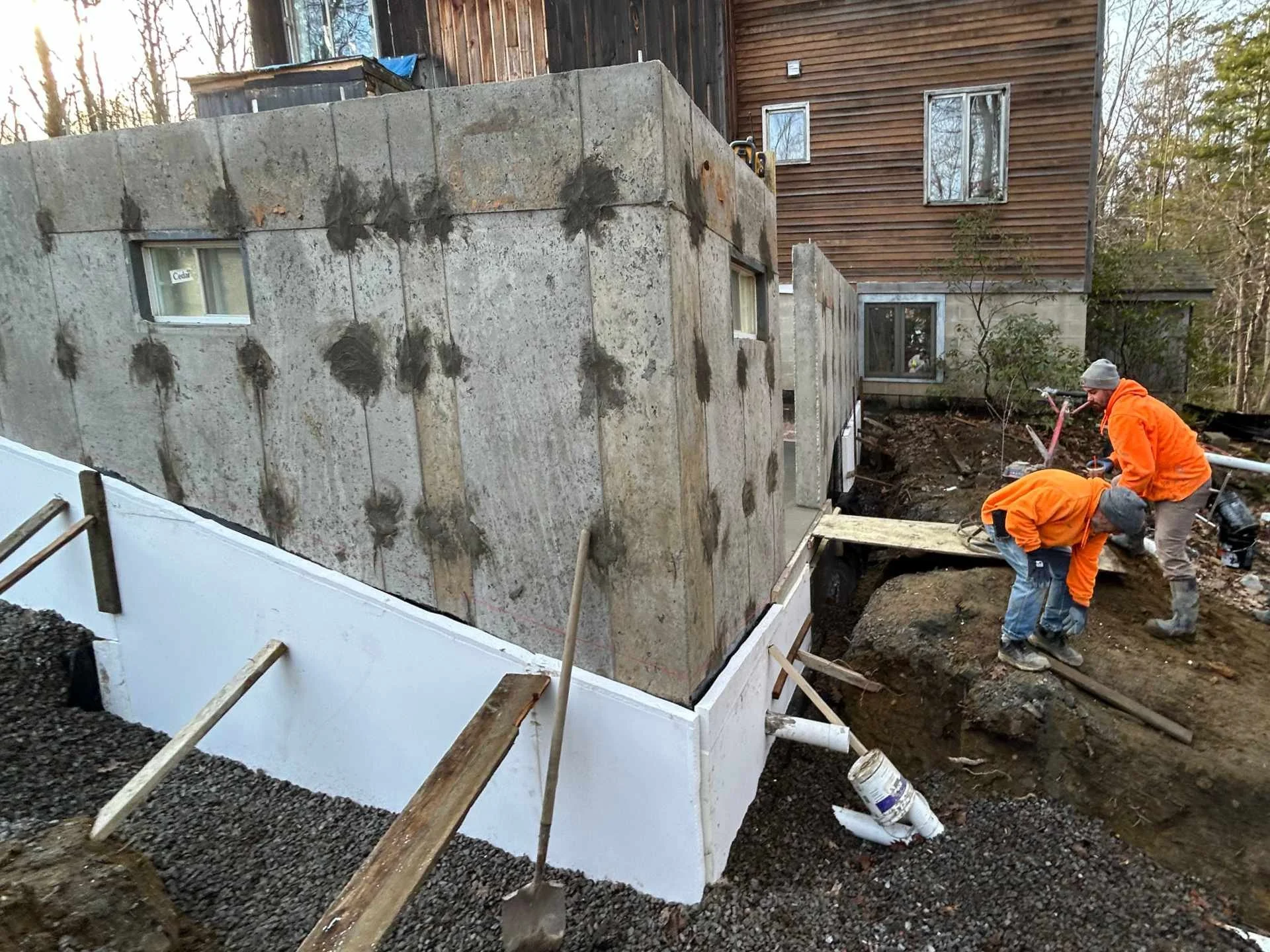 Construction workers placing insulation around concrete foundation walls around a house in a wooded area.