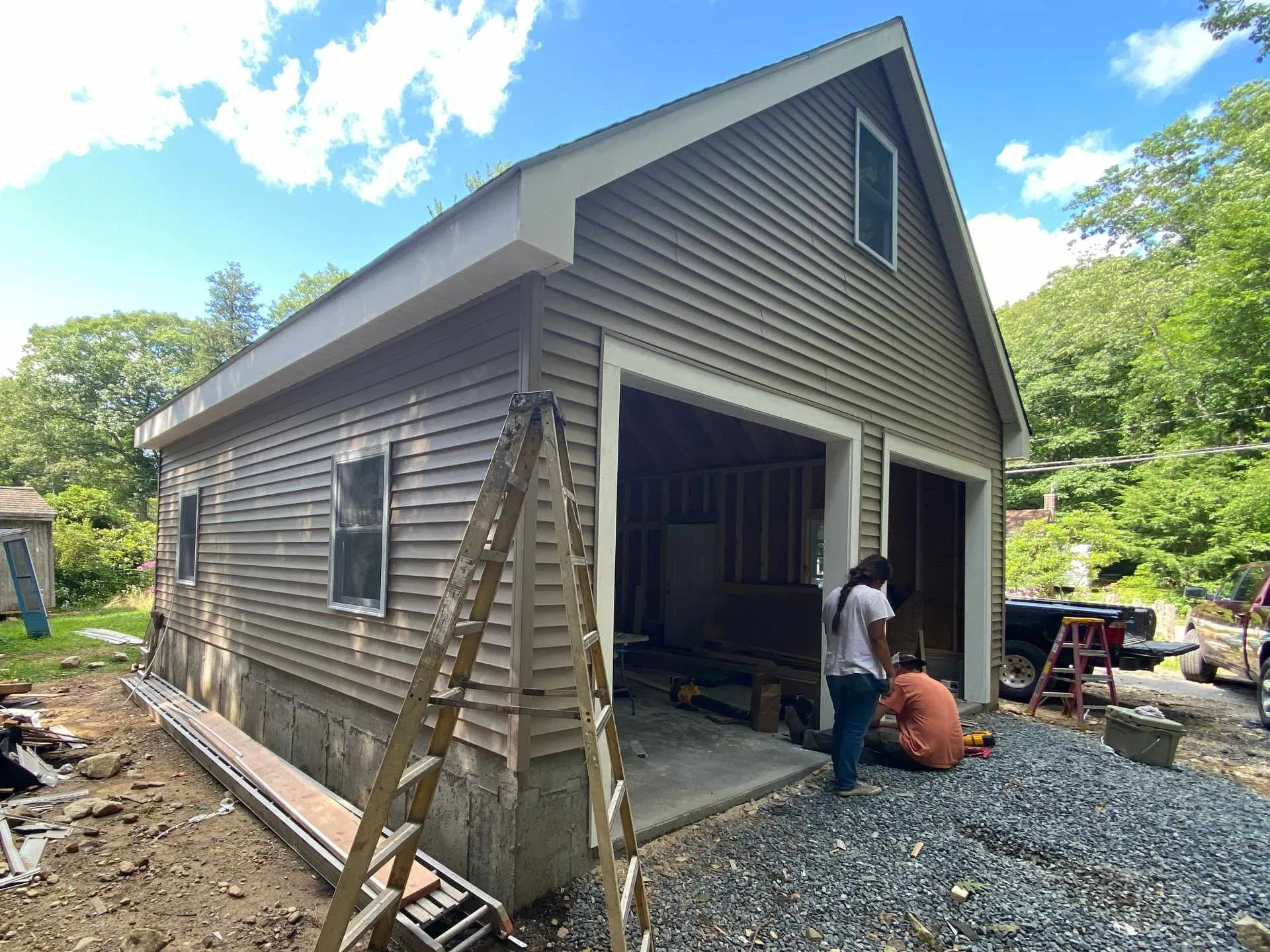 Construction workers building a house, siding being installed, ladders and tools around, an open garage or shed, trees and a blue sky in the background.
