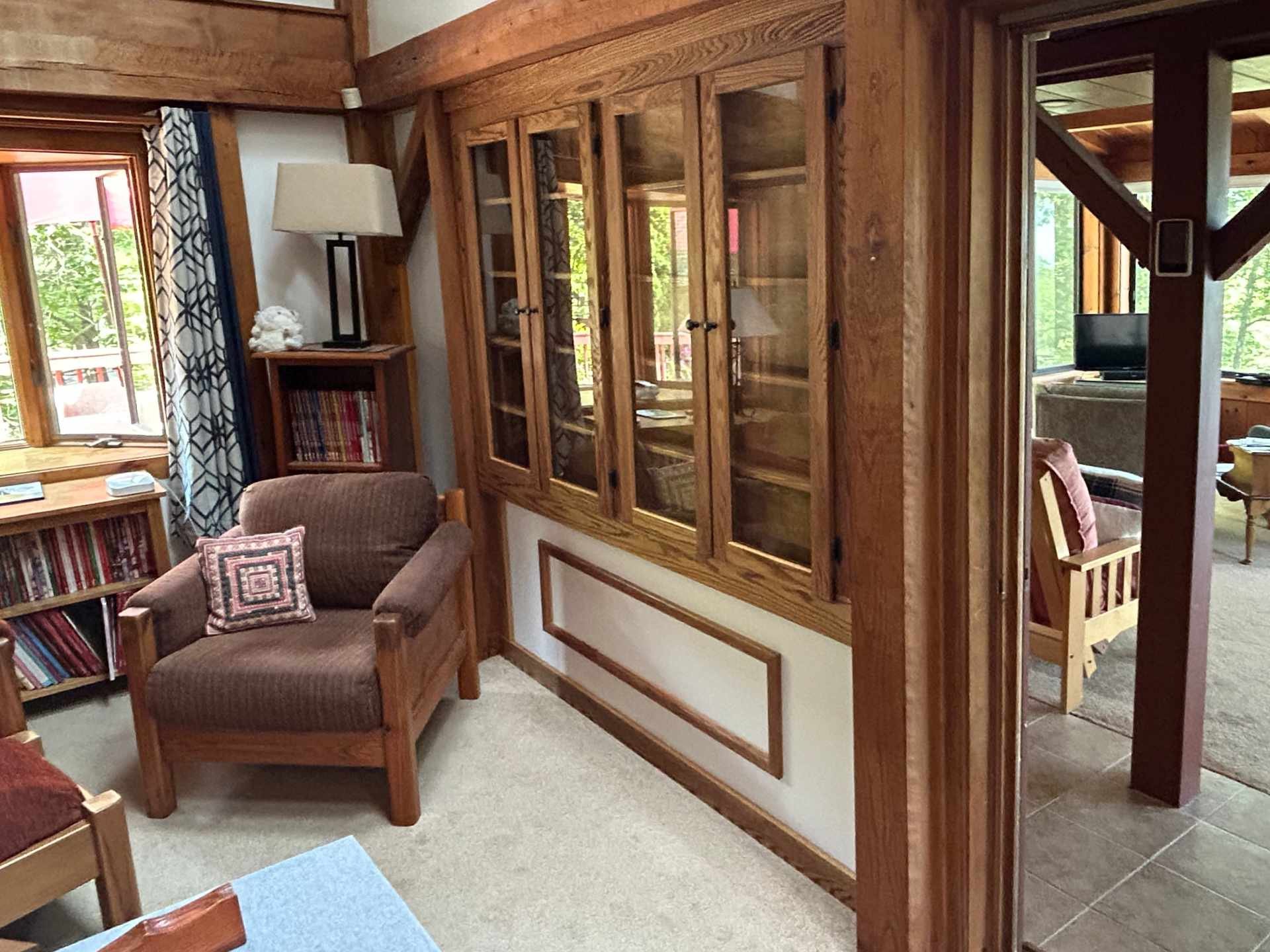 Living room with wooden oak features including a custom built in bookcase and trim with custom glass doors. There are windows with patterned curtains and an adjoining addition room visible through a wooden sliding door.