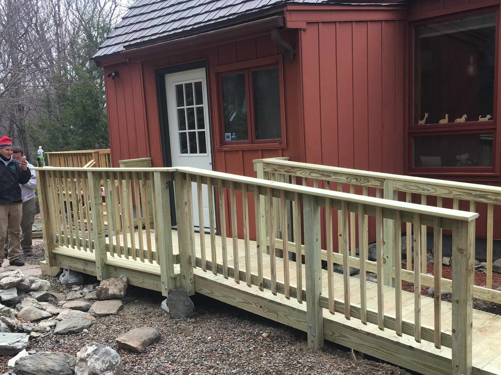 A newly built wooden ramp with a railing in front of a red house with a white door and window, surrounded by scattered rocks and trees.