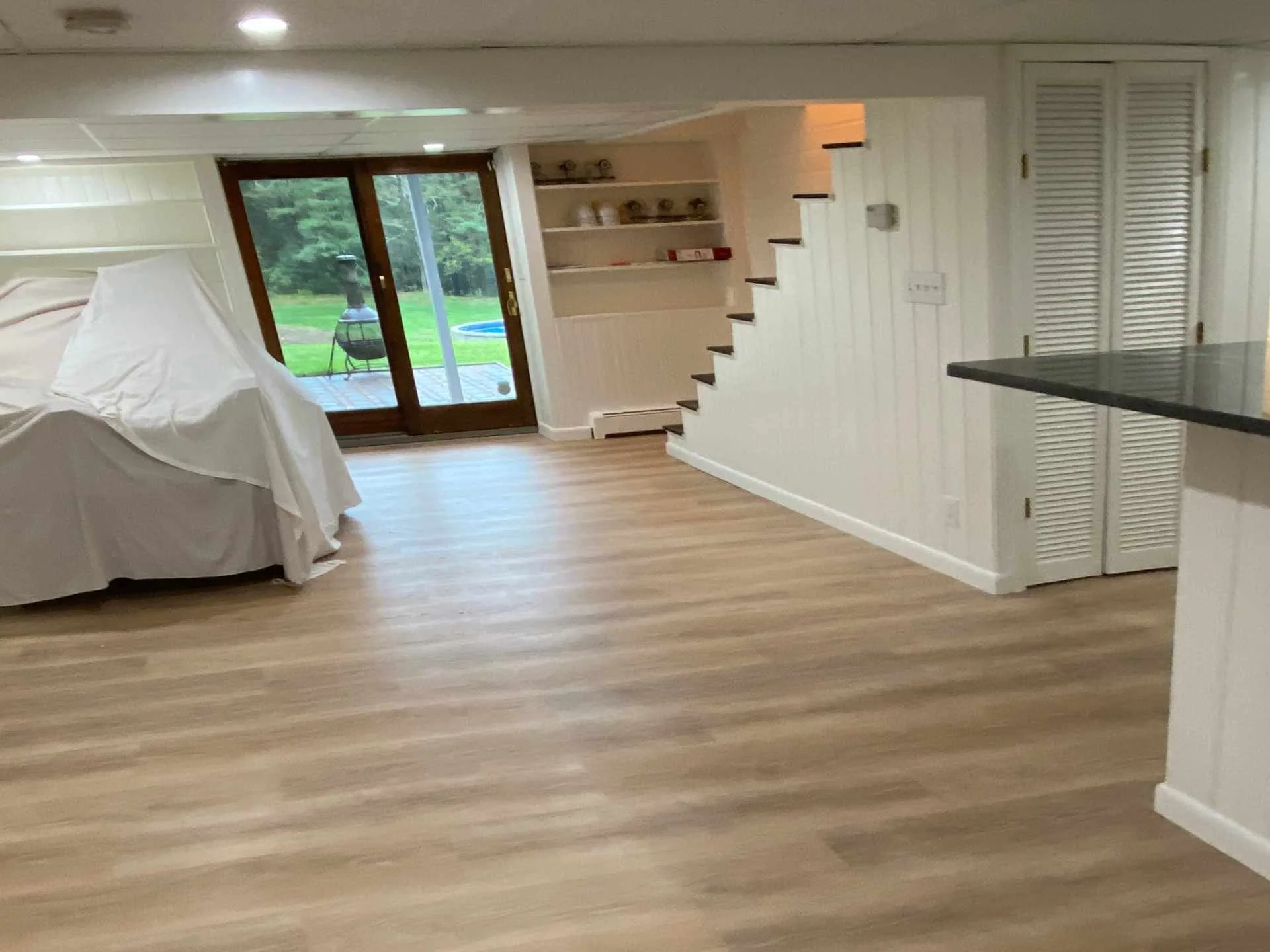 Living room with glass sliding door leading to a backyard, staircase along right wall, white paneled walls, a covered piece of furniture, and wooden flooring.