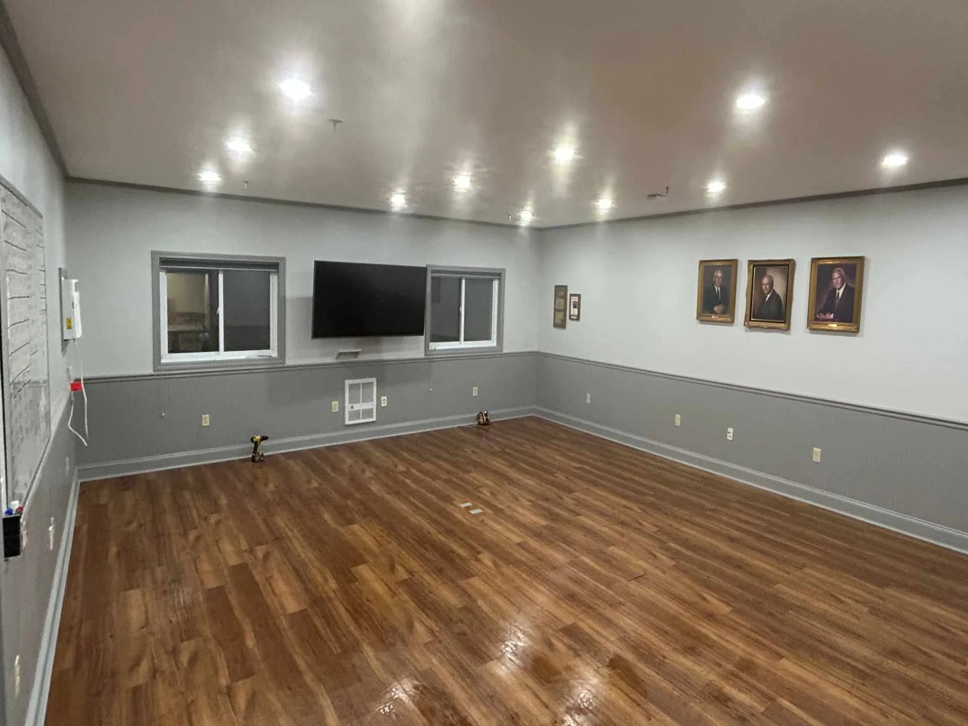 Empty conference room at a commercial property with brown wooden flooring, white walls, LED recessed ceiling lights, framed portraits on the wall, windows, and a television mounted on the wall.