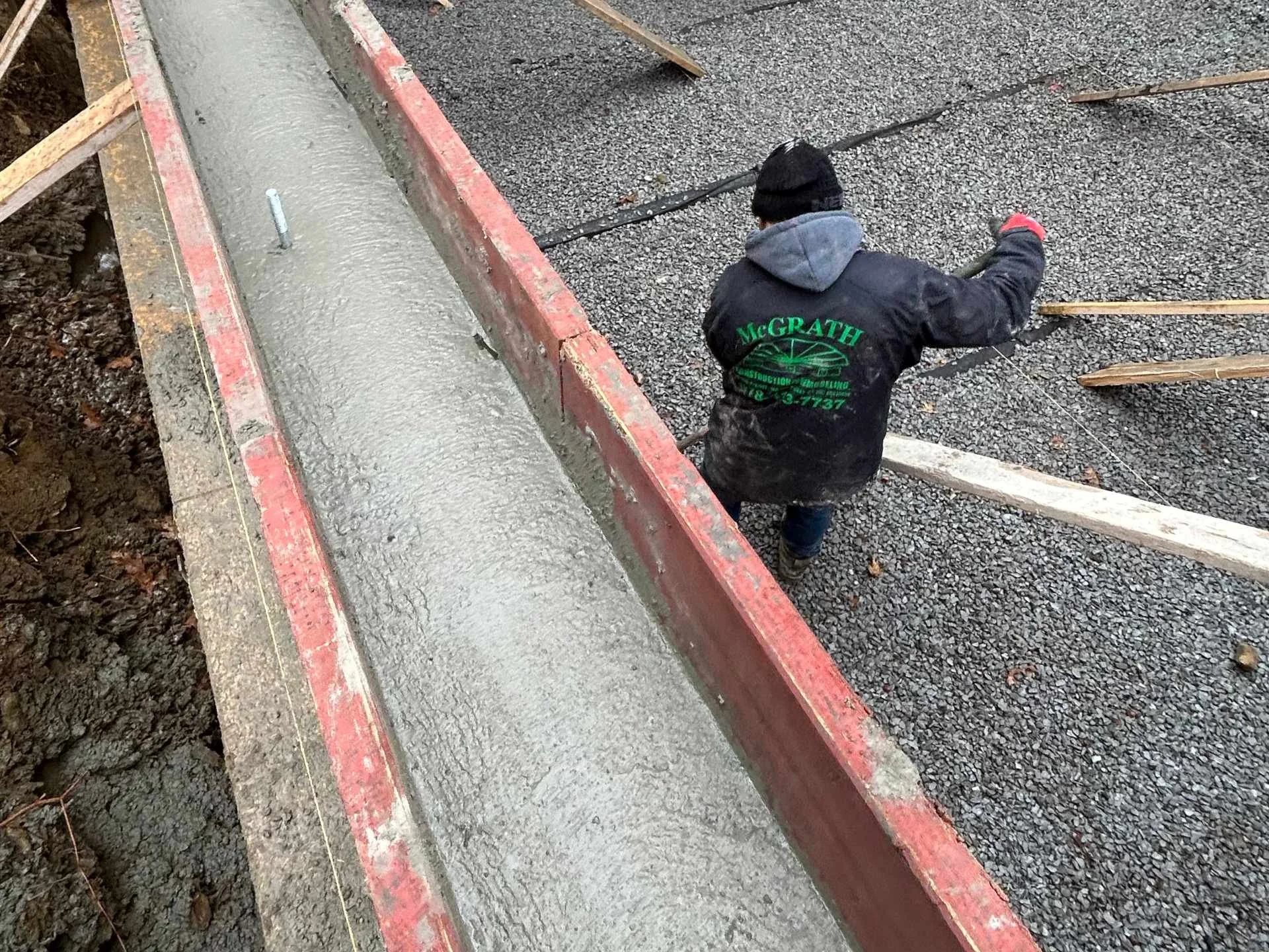 A construction worker wearing a black MCGRATH jacket with green lettering is leveling crushed stone next to a freshly poured concrete foundation wall. The foundation wall is framed with wooden and metal forms.