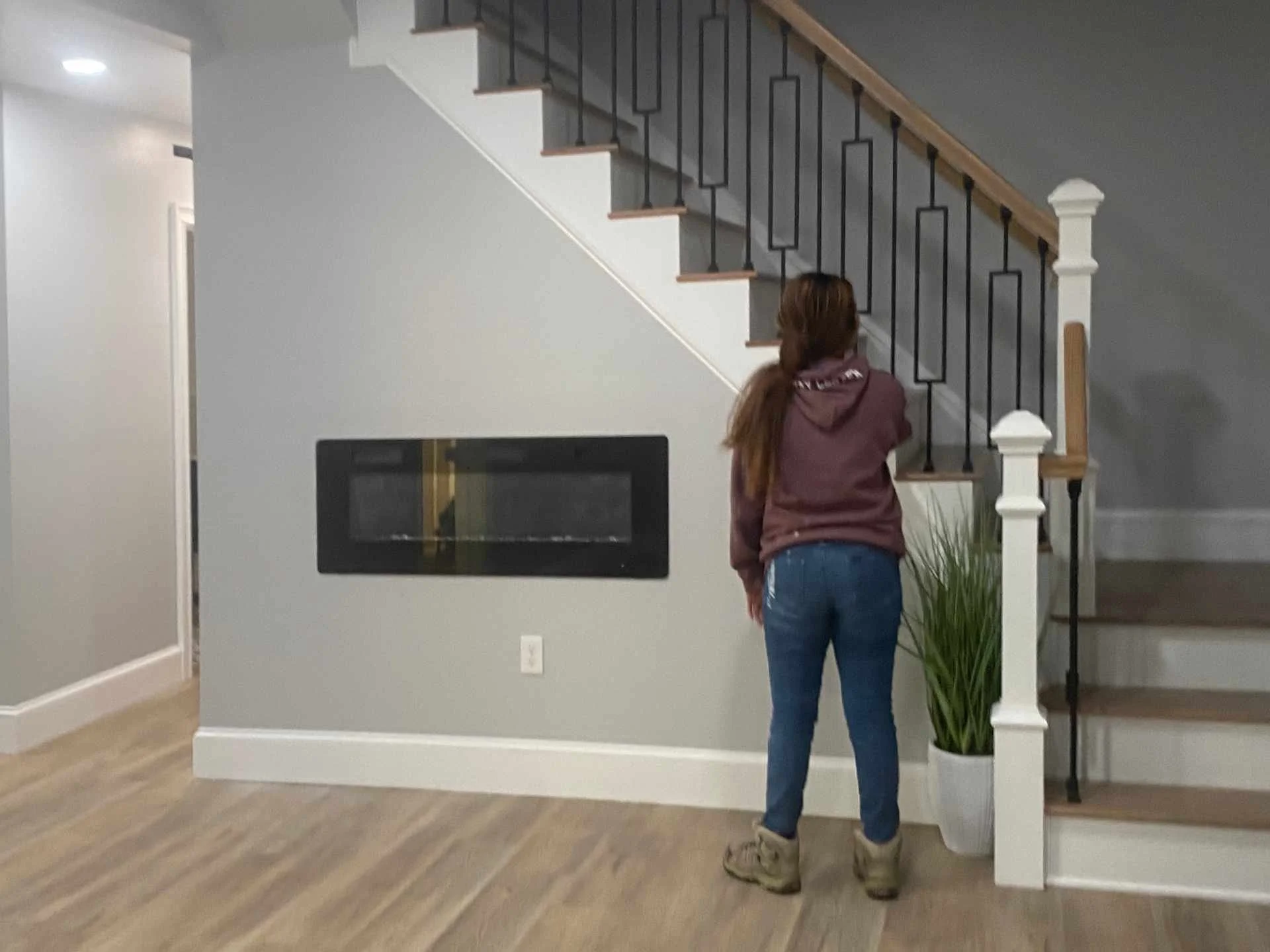 A woman with long black hair, wearing a maroon hoodie, blue jeans, and tan boots, standing inside a house with a new finished basement near a staircase with black railings and white decorative posts. There is a new gas insert.