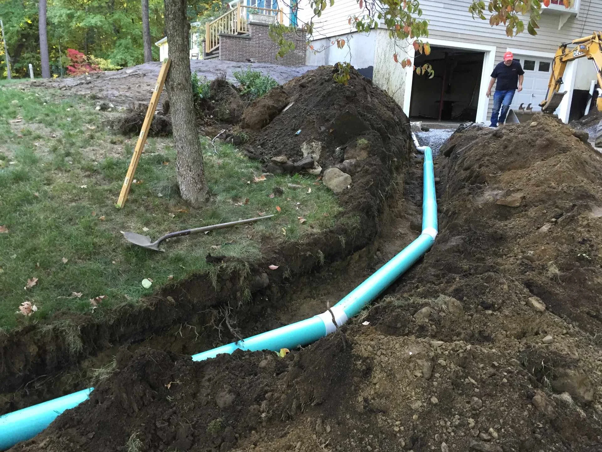 Dig site in a yard with a large trench and a light blue pipe, with a person walking near a small excavator, trees, grass, and a house in the background.