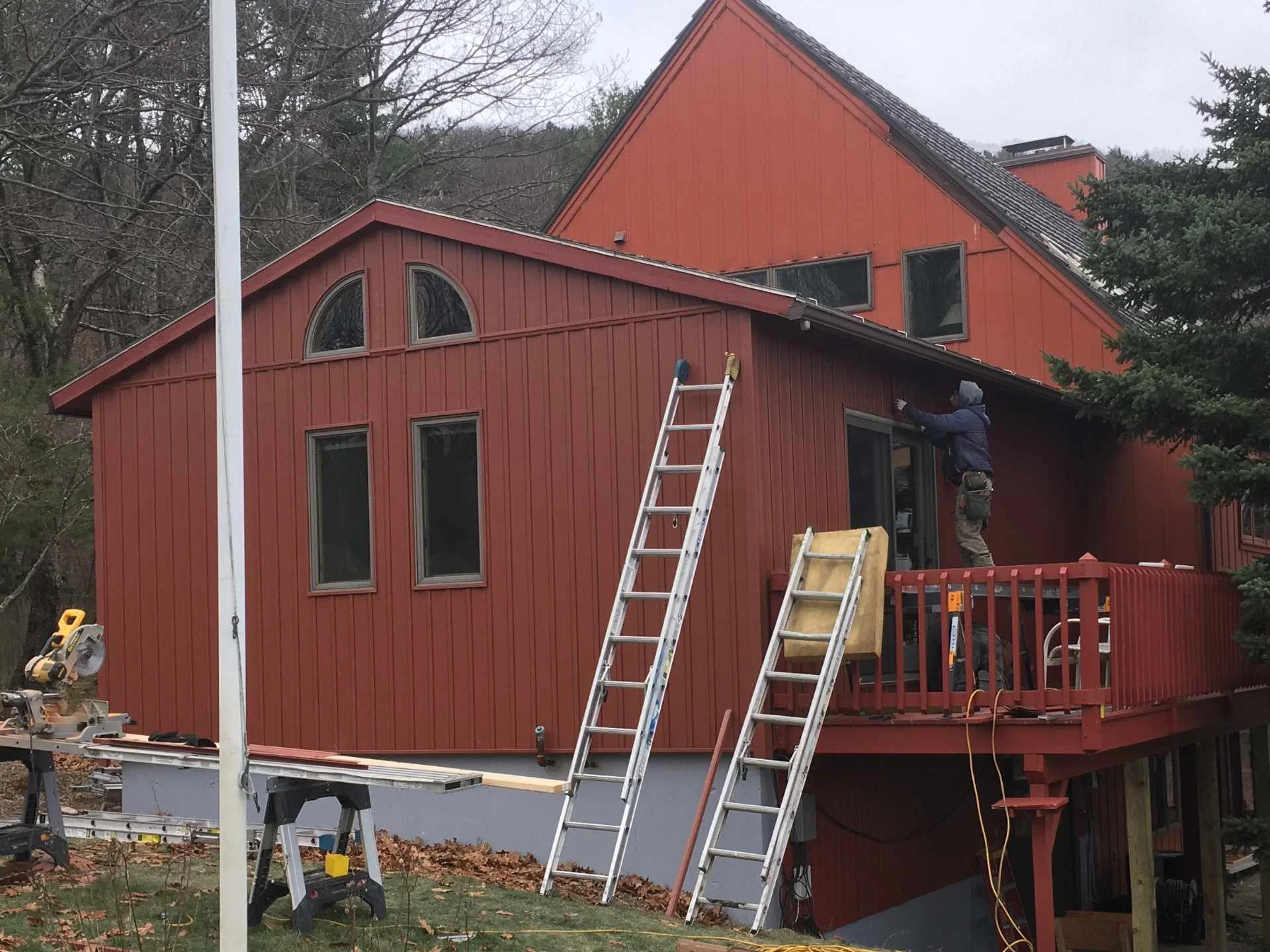 A person working on the exterior of a red house on Wachusett Mountain with a steeply pitched roof. Tools and ladders are set up around the house, and a large tree is nearby.