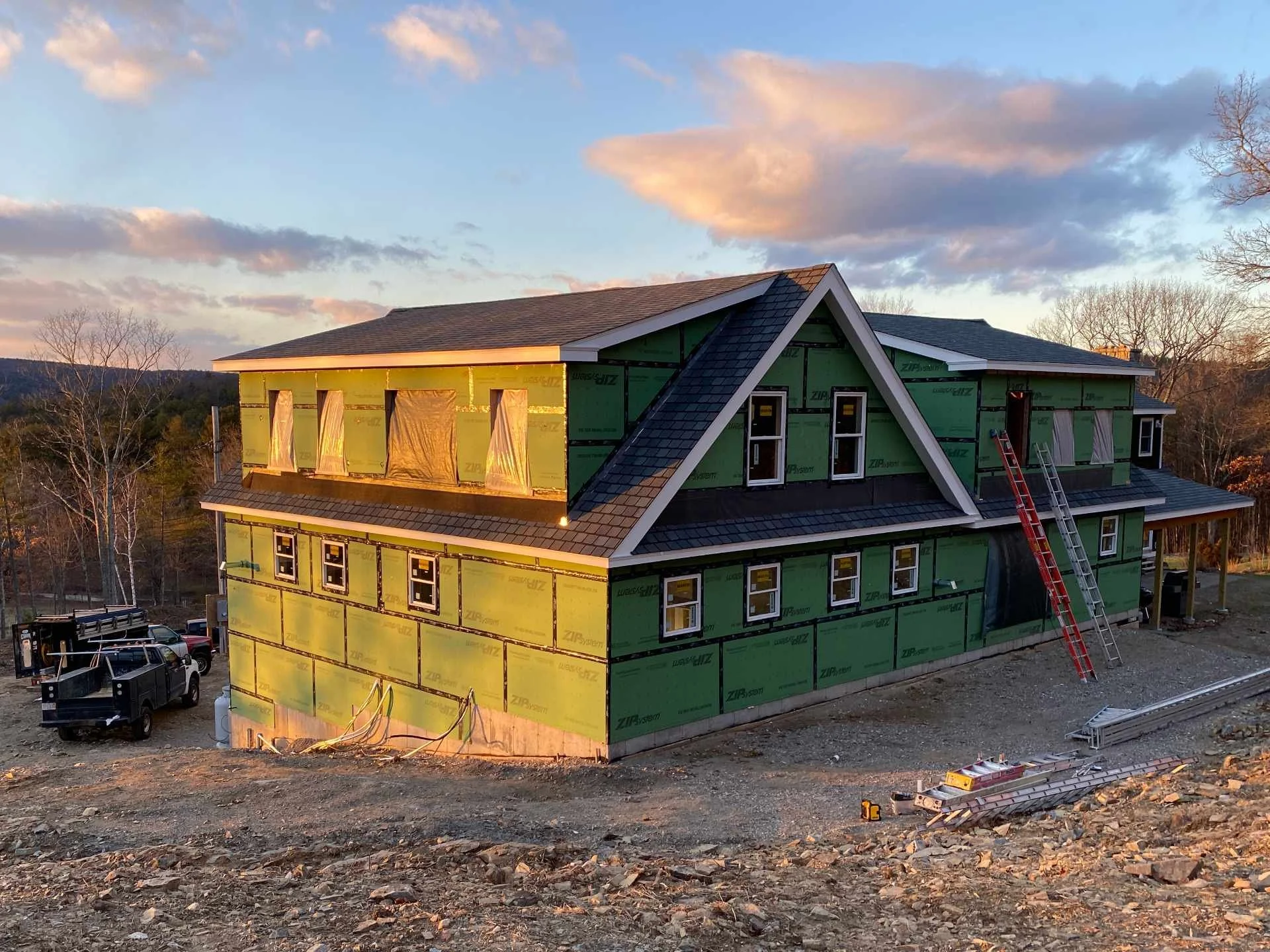 A house under construction during sunset, partially covered with green Zip System sheathing, with a roof and multiple windows installed and ladders and construction equipment visible around the building.