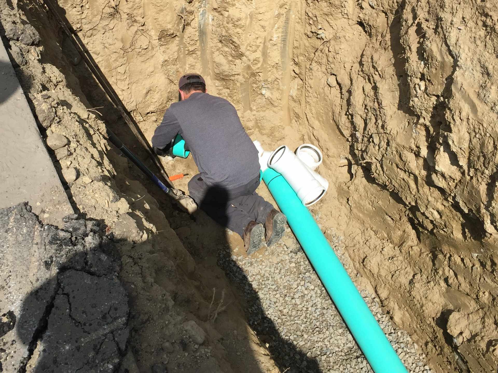 A man working on installing a blue pipe in an excavated trench with rocky soil walls.