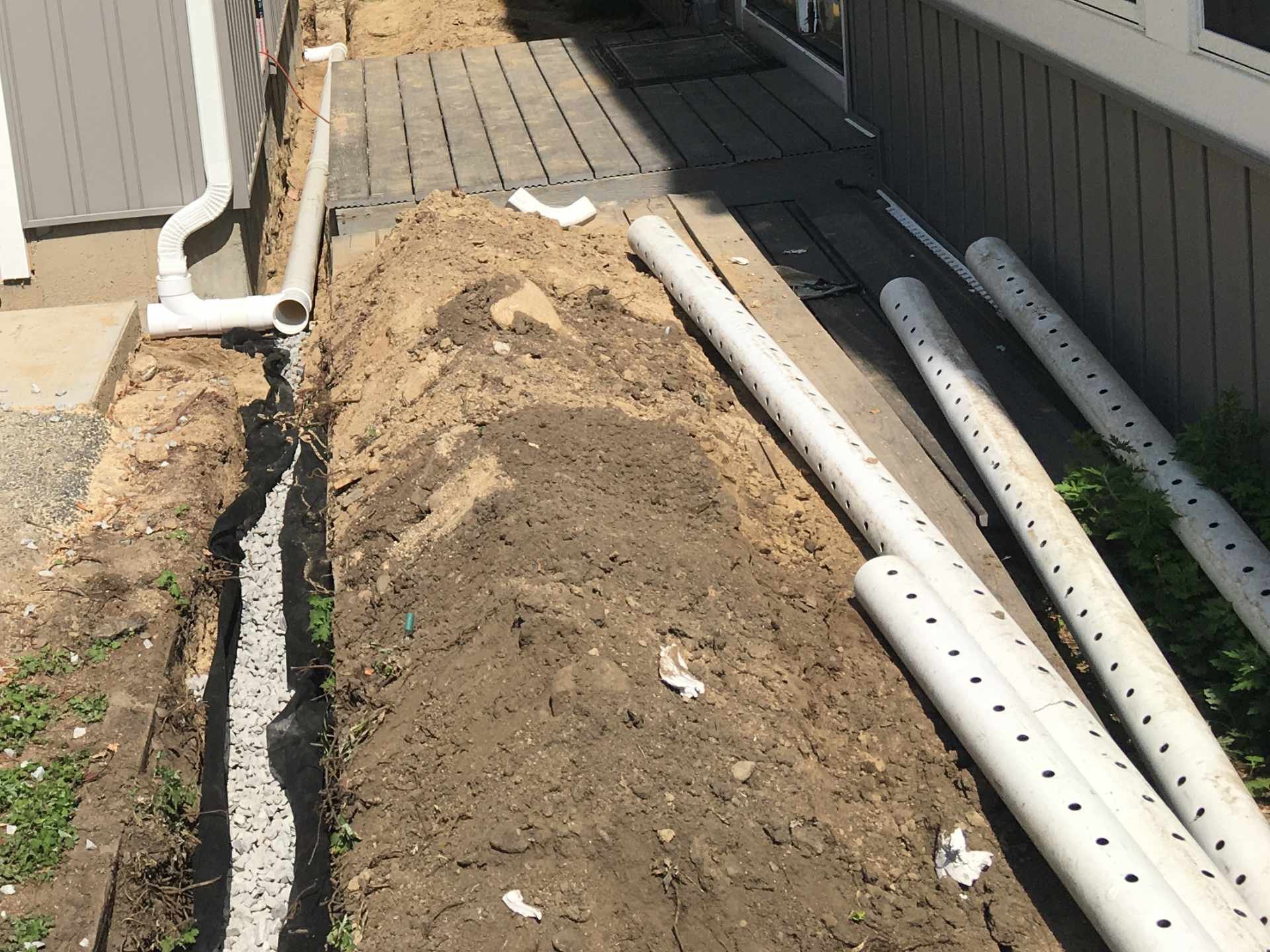 Construction site with exposed pipes, a black cable, some dirt, and a small area of grass, with a building wall and a door in the background.