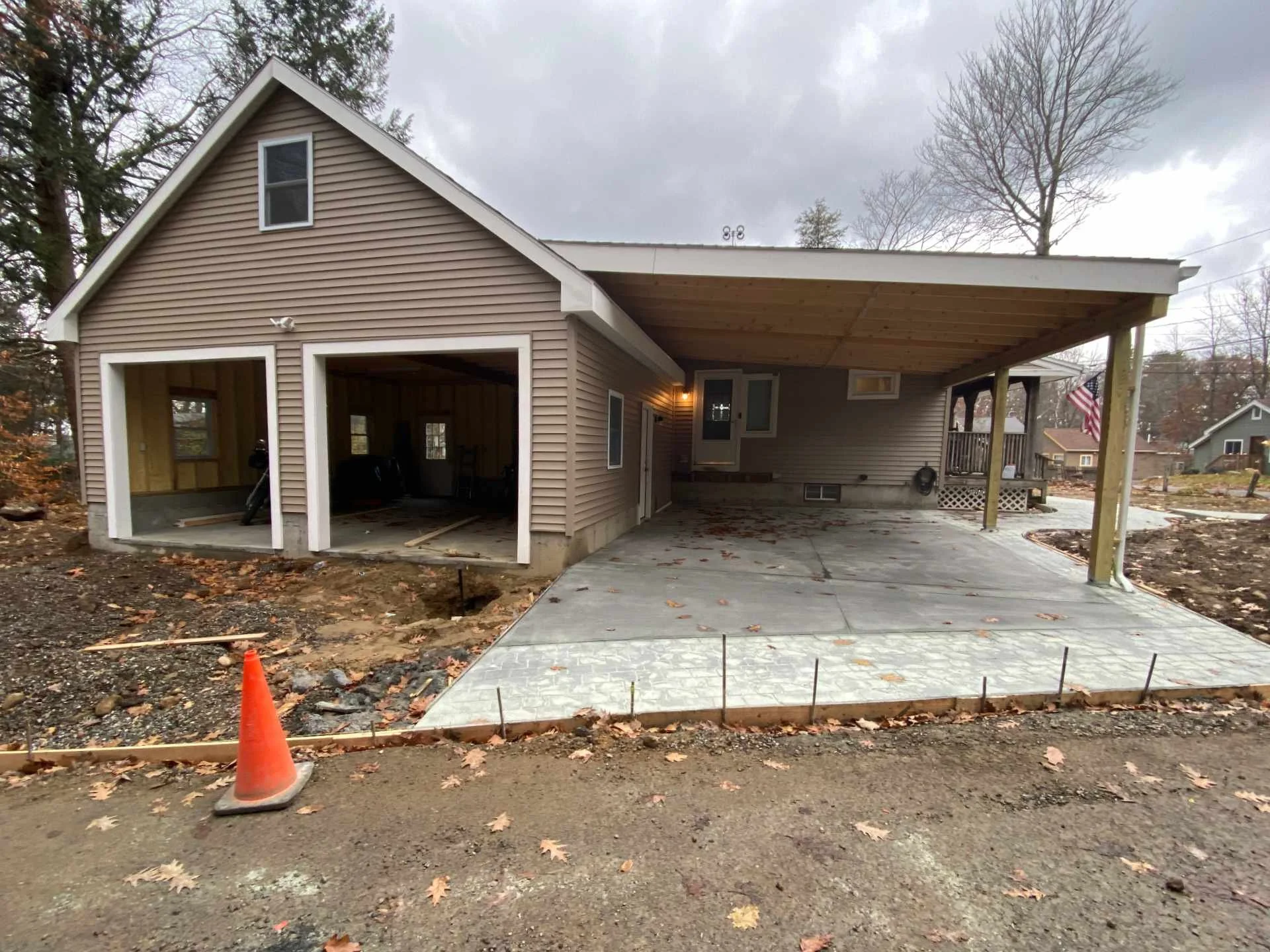 A house under construction with a garage and a porch. The driveway is concrete, and there is a small construction cone in the foreground. The exterior walls are partially finished with siding, and the garage door is open, showing the unfinished inter