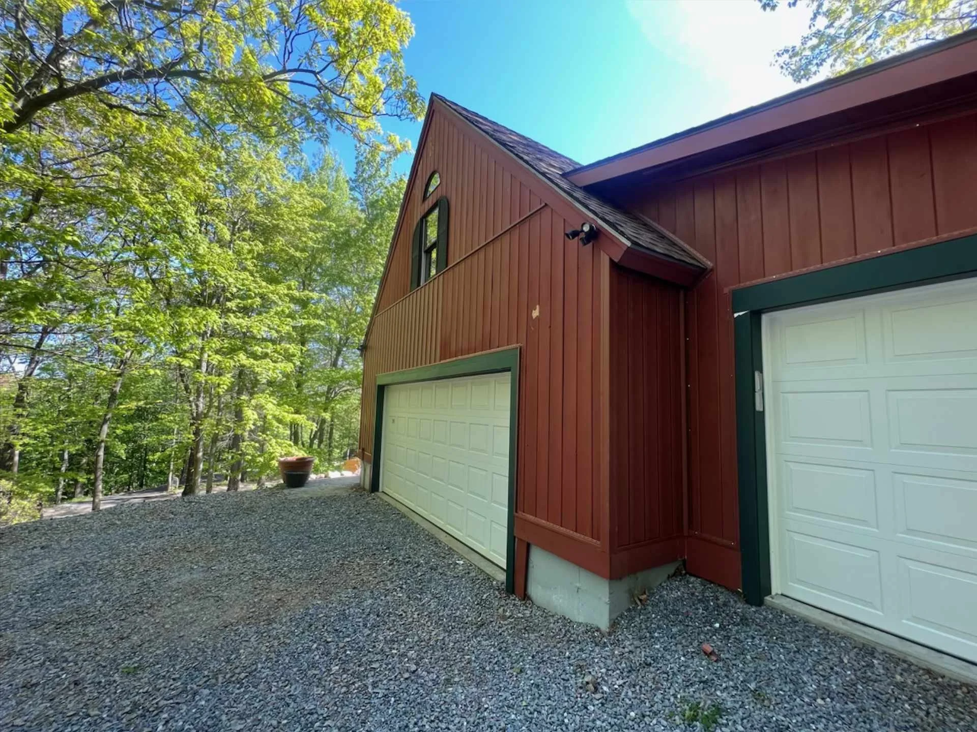 A red barn-style garage with green trim and two garage doors, surrounded by trees and a gravel driveway, under a blue sky.