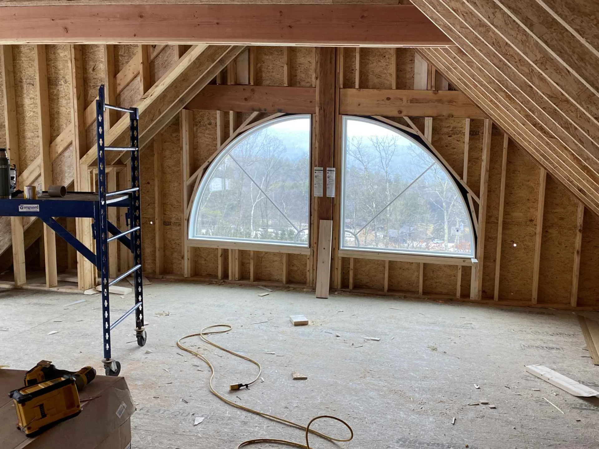 Interior of a house under construction with exposed wooden framing, a large arched window, a metal scaffold, and construction tools on the floor.