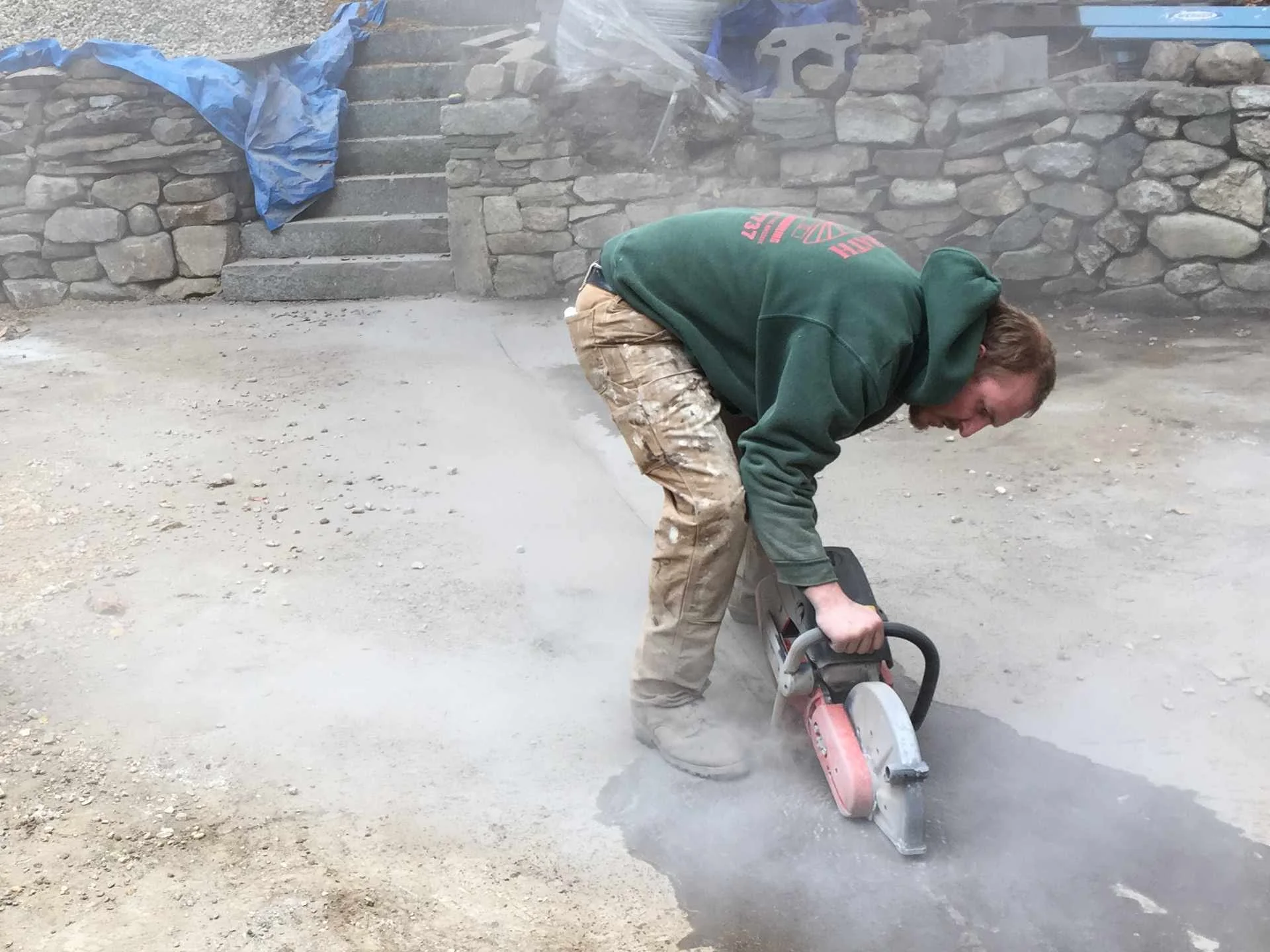 A man using a power saw to cut concrete on a construction site, with stone steps and a stone wall in the background.