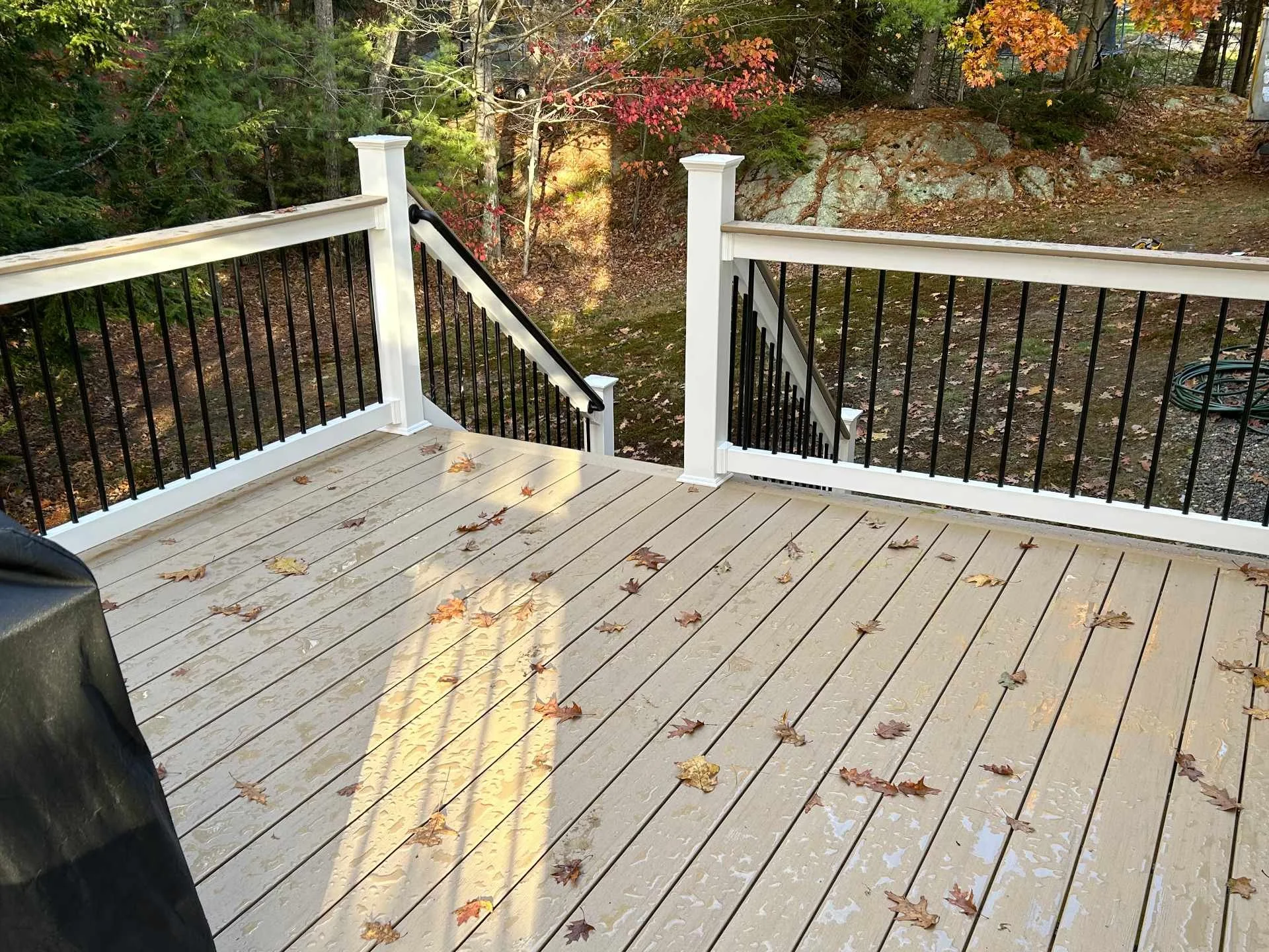 A wooden deck with fallen autumn leaves, overlooking a wooded area with trees displaying fall foliage and rocky terrain.