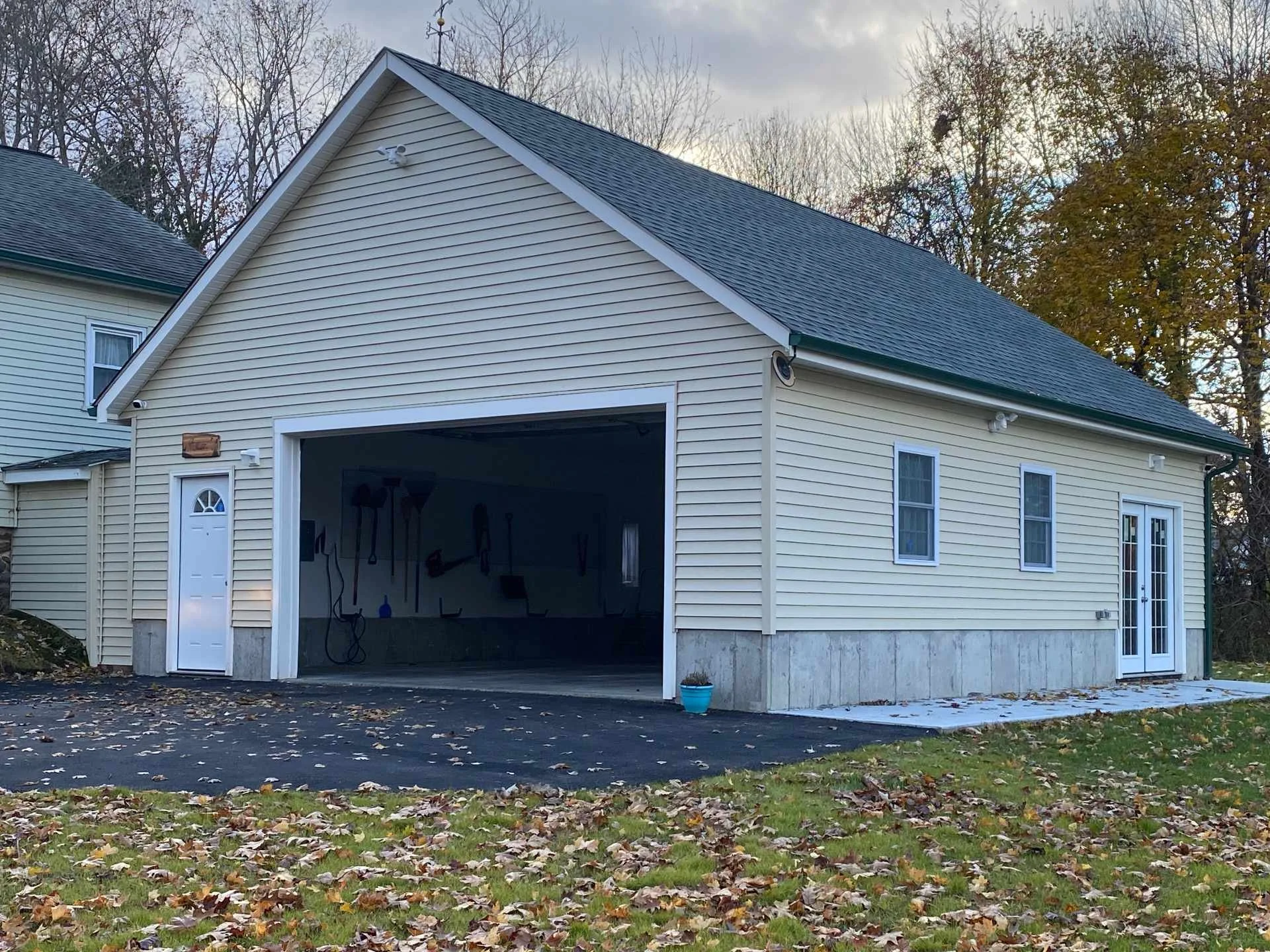 A yellow two-story house with a garage, white trim, and a pitched roof, situated in a yard with fallen leaves and a patch of snow, with a background of leafless trees and overcast sky.