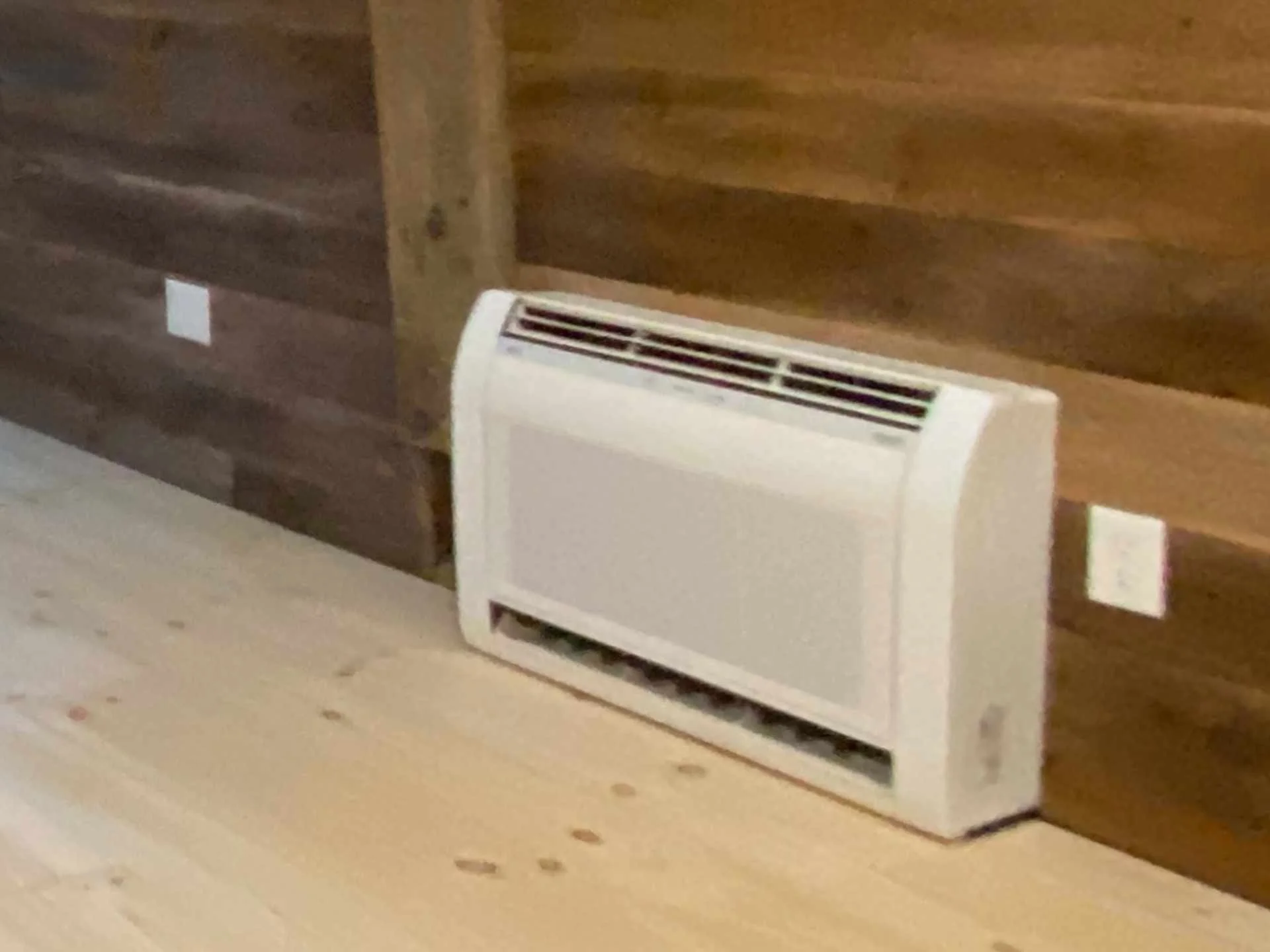 White floor-mounted Fujitsu air conditioning/heating unit placed on a light-colored floor in front of a wooden wall with horizontal planks.