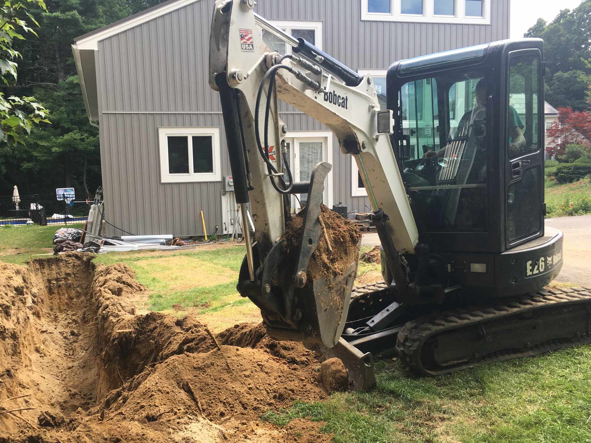 A small excavator digging a foundation trench for a new garage in a backyard of a residential house with a gray exterior, surrounded by trees and a fence.