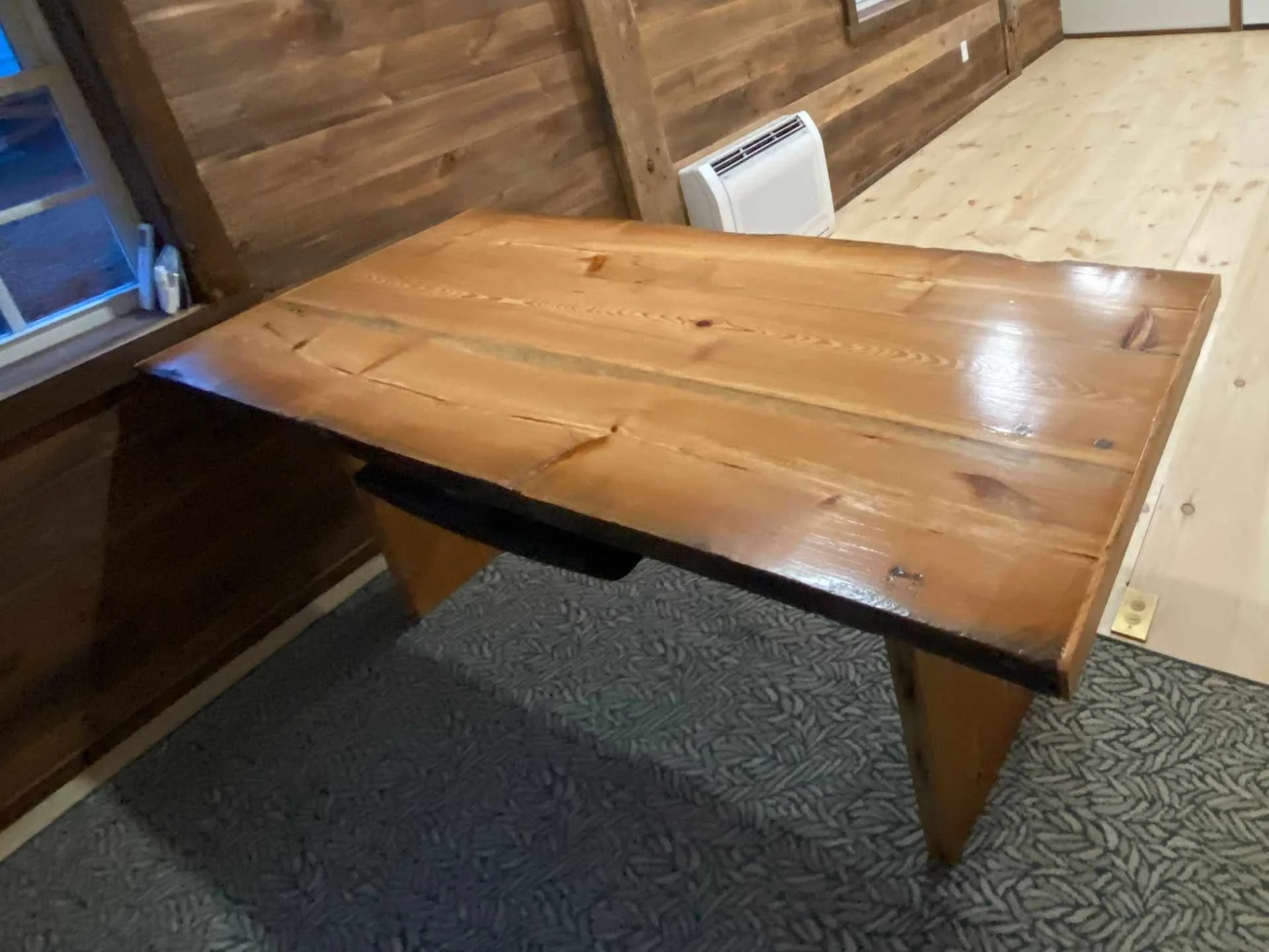 A custom reclaimed heart pine wooden dining table with a natural finish in a room with wood-paneled walls and pine floor, near a window with blinds and household items.