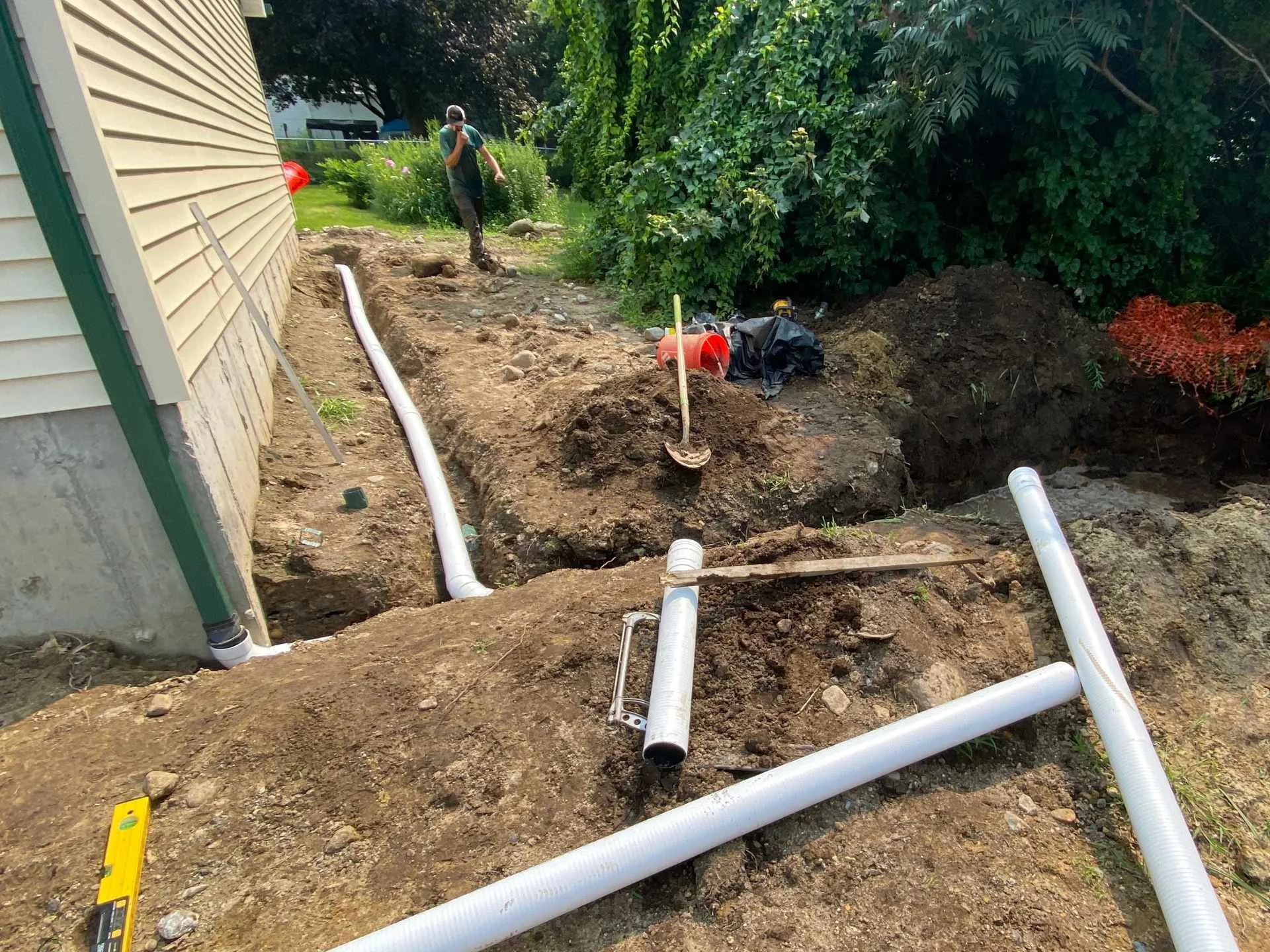 A construction worker stands in a trench next to the side of a house, installing underground pipes with tools, construction materials, and dirt around.