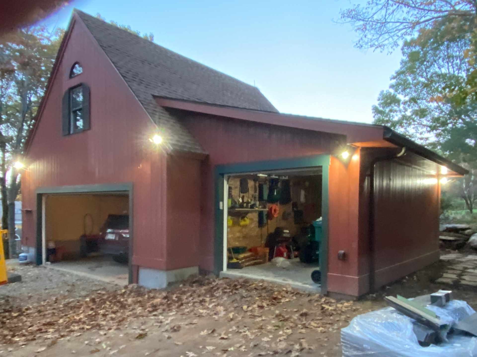 A garage in a house with an open door, revealing tools and storage inside. The house has a red exterior with teal framing around the garage door. There are trees with fallen leaves on the ground, and exterior lights are on, indicating early evening o