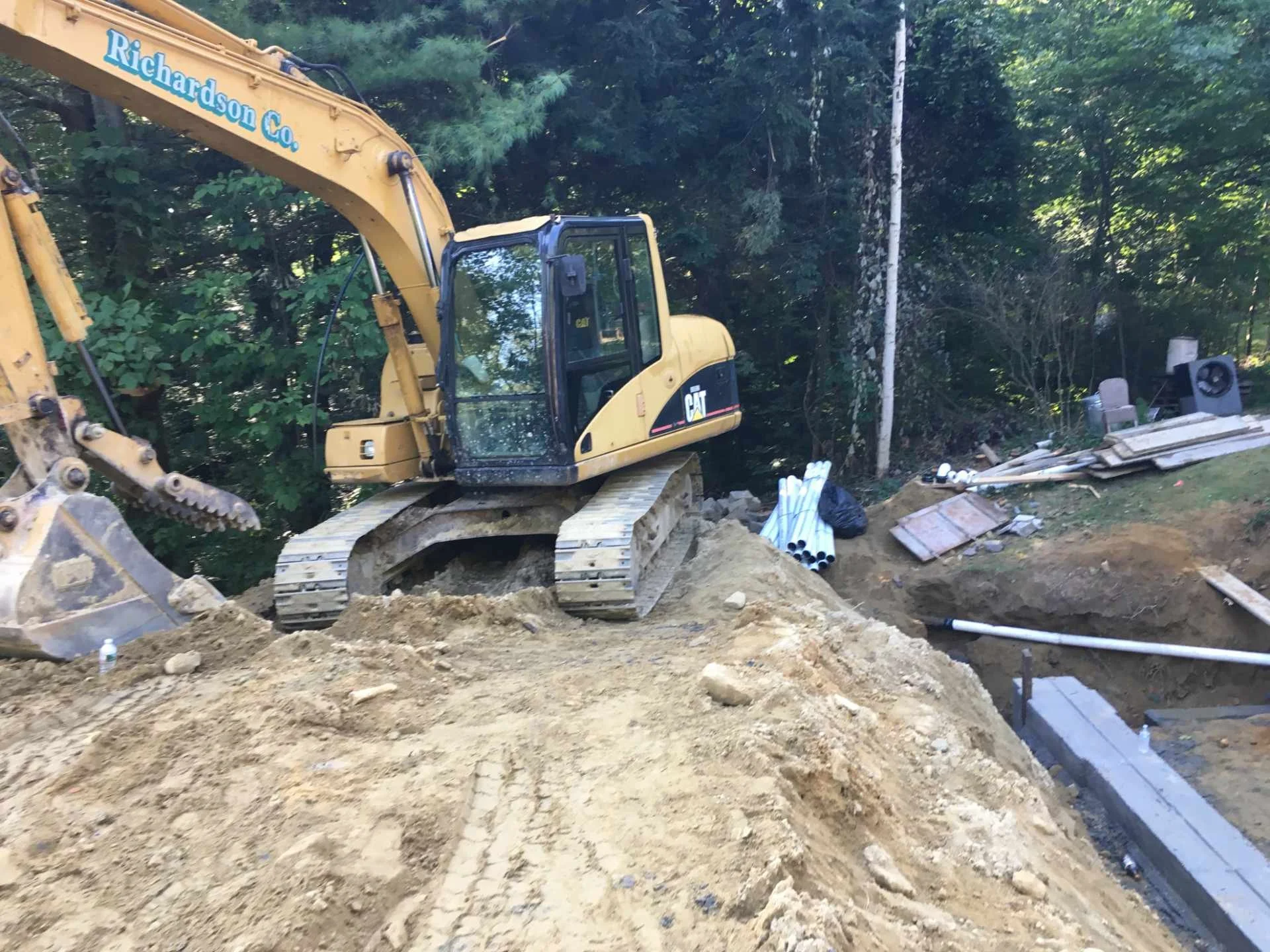 A yellow Caterpillar excavator working on a construction site surrounded by dirt, debris, pipes, and trees in the background.