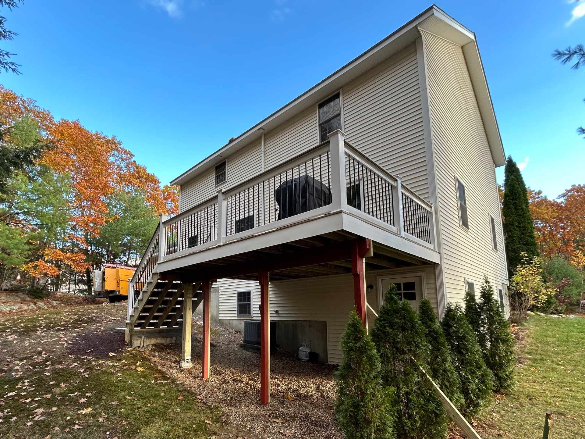 Backyard view of a two-story house with a large wooden deck, stairs, and white vinyl railing with black aluminum ballusters, and a covered grill, surrounded by autumn trees and a clear blue sky.
