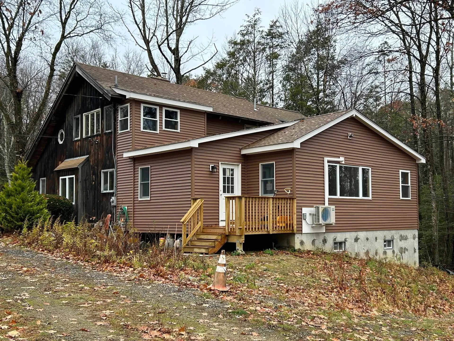 A two-story house with brown siding and some blackened wood on the upper left section, surrounded by leafless trees, with a small wooden porch staircase and an air conditioning unit outside on the lower right.