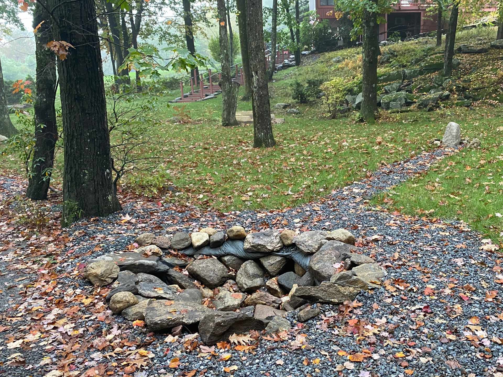 A small stone bridge over a dry creek bed in a wooded yard, fallen leaves on the ground, trees and a house visible in the background.