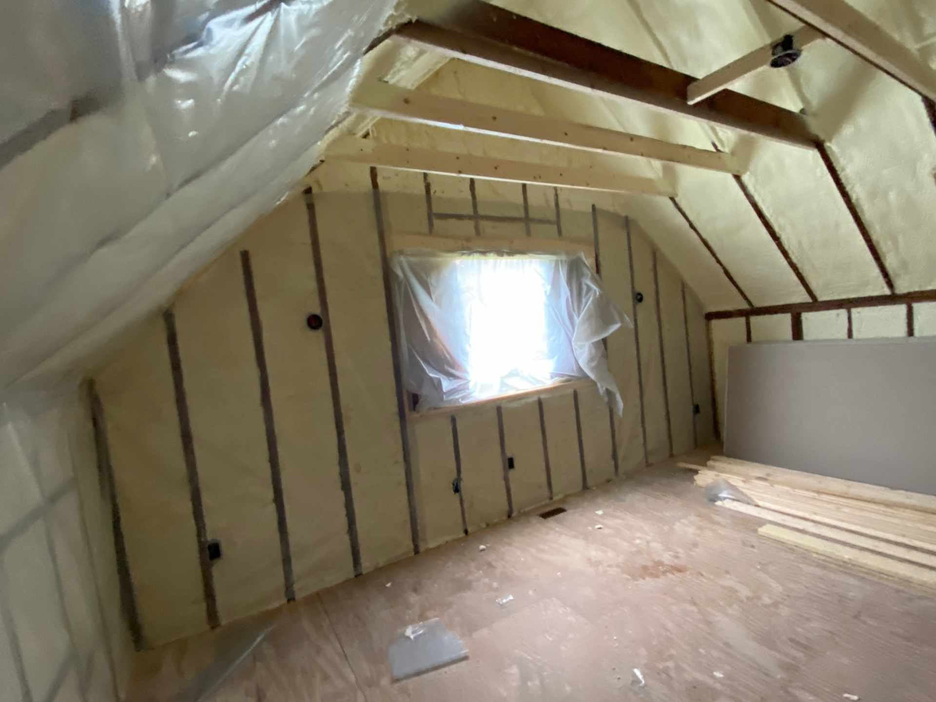 An attic or unfinished room with exposed wooden framing, a small window covered with plastic, and spray foam insulation already installed on the walls and ceiling.
