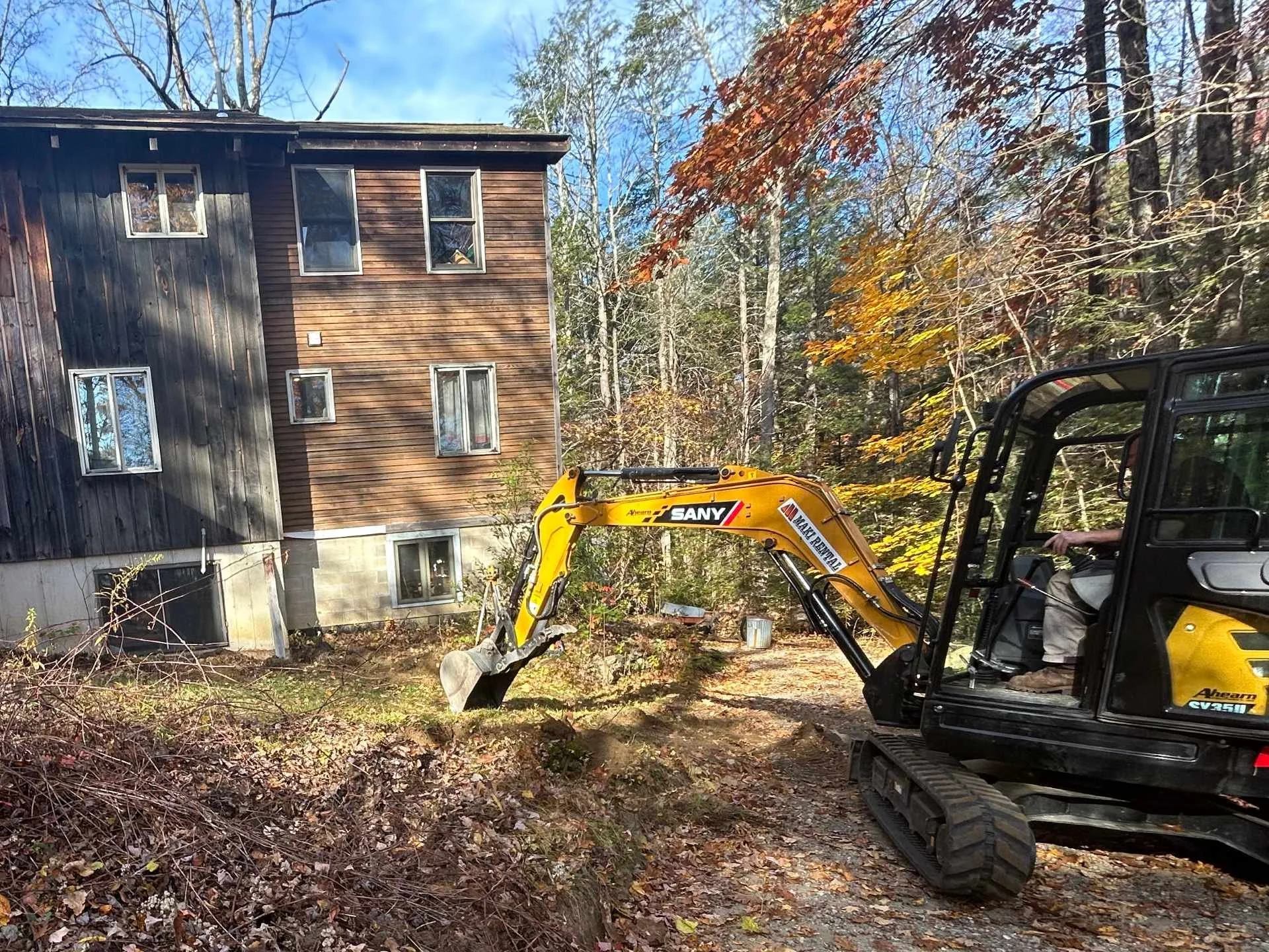 Construction worker operating a small yellow excavator near a multi-story wooden house in a wooded area with autumn leaves.