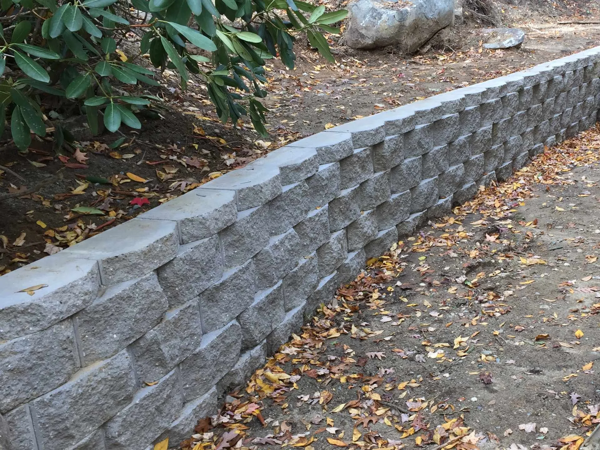 A stone retaining wall made of rectangular gray stones with a rough texture, separating a dirt area with fallen autumn leaves from a garden bed with green leafy plants and rocks.