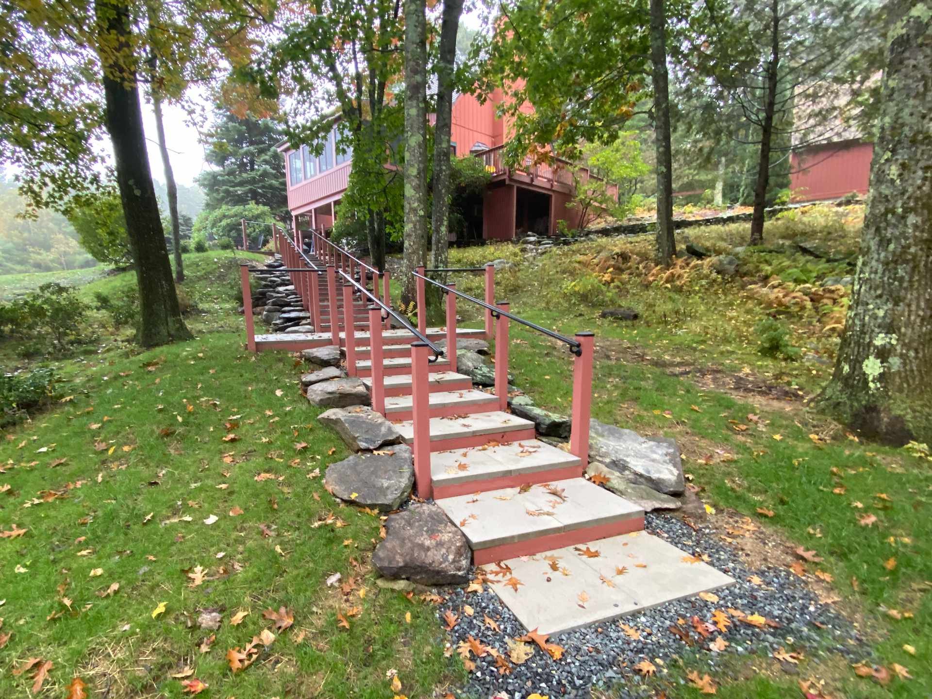 A wooden frame staircase with large patio pavers and metal railings leading up a grassy hill with trees, rocks, and a red house in the background.