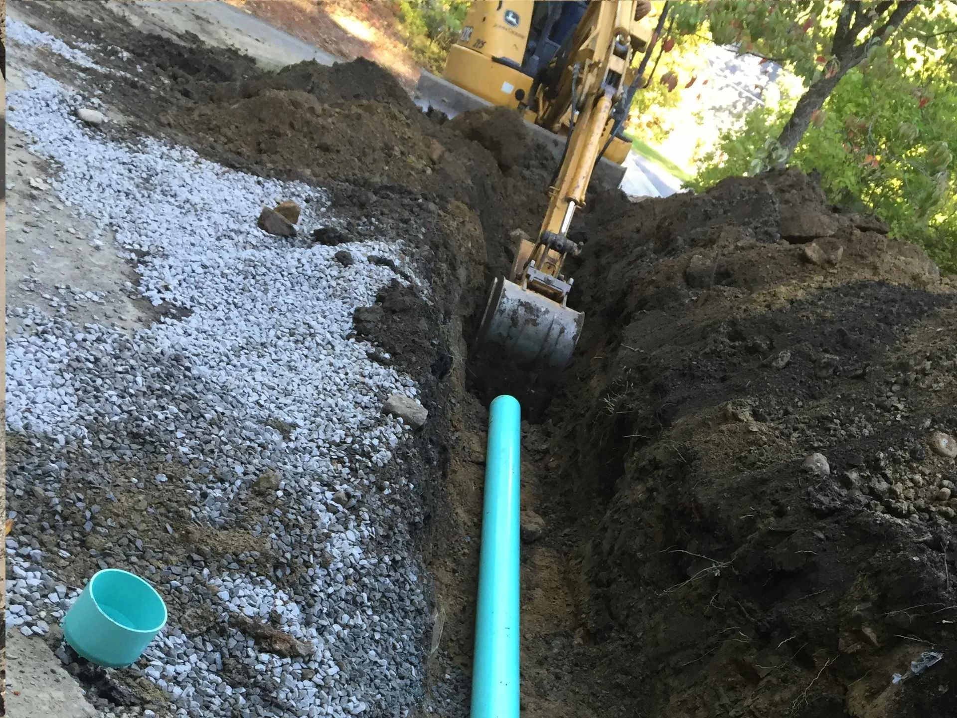 Excavation site with a yellow backhoe digging a trench in the soil, near a tree with green leaves; a blue pipe is laid in the trench, and a blue bucket is on the ground nearby.