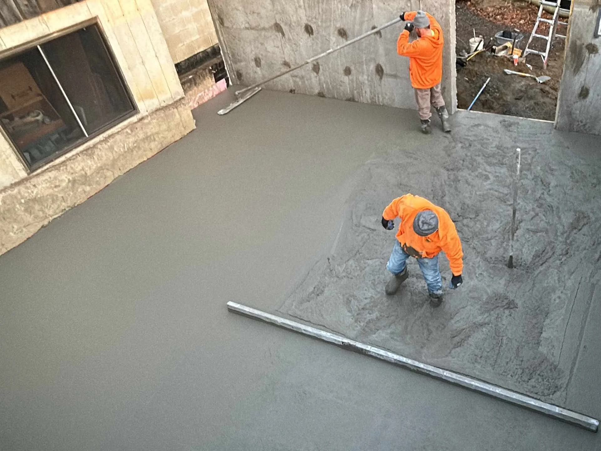Construction workers in orange jackets leveling wet concrete on a construction site.