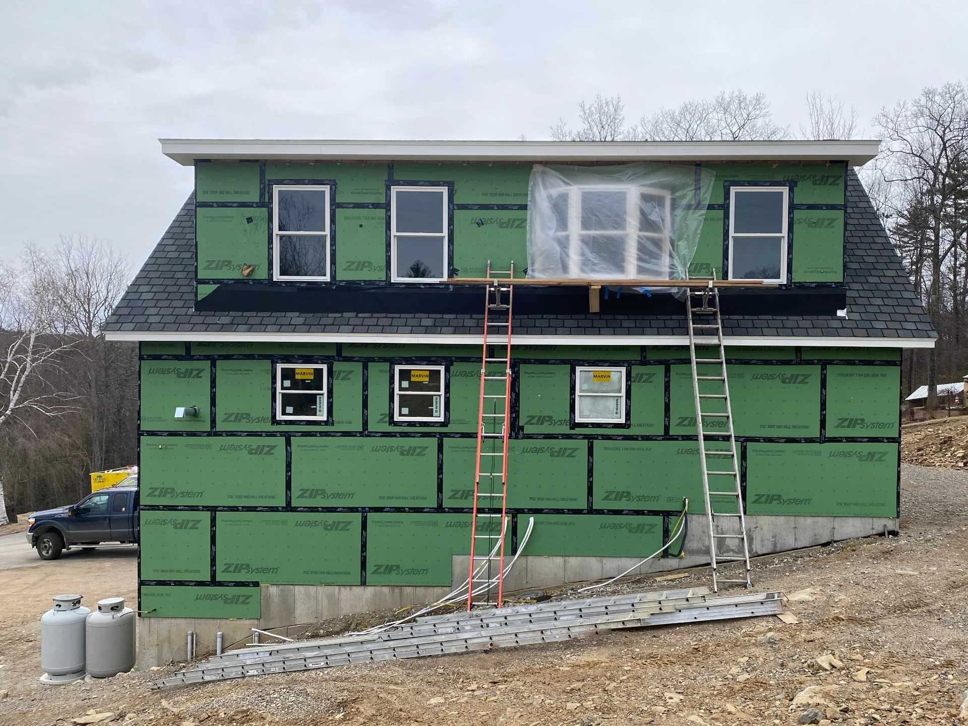 Two-story house under construction with green sheathing and black trim, scaffolding and ladders at the front, roof partially finished, and vehicles and propane tanks in the yard.