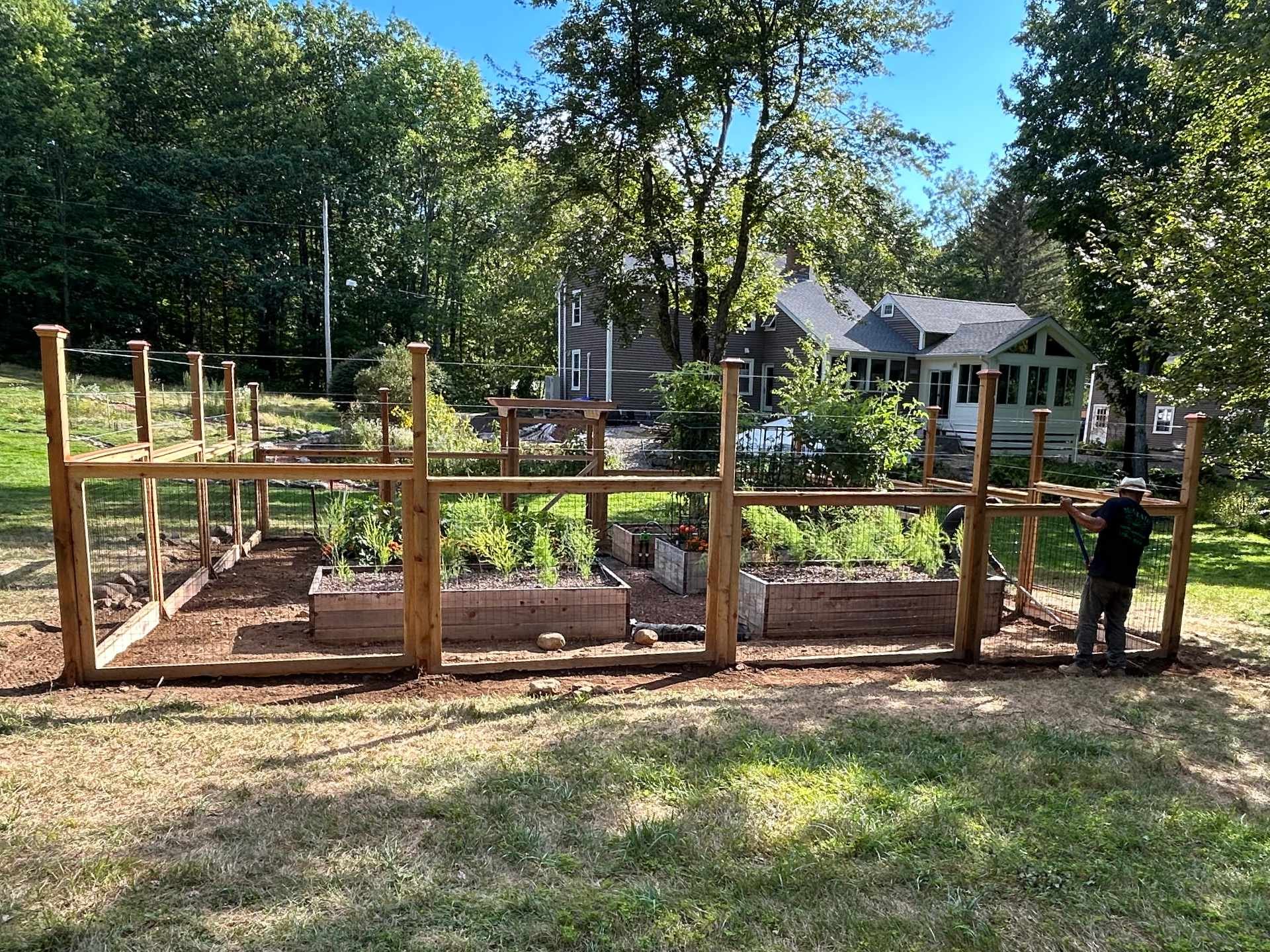 The image shows a backyard garden enclosed with a wooden and wire fence with garden beds containing plants. A person wearing a hat is working in the garden. In the background, there is a house with gray siding and multiple windows, surrounded by tall