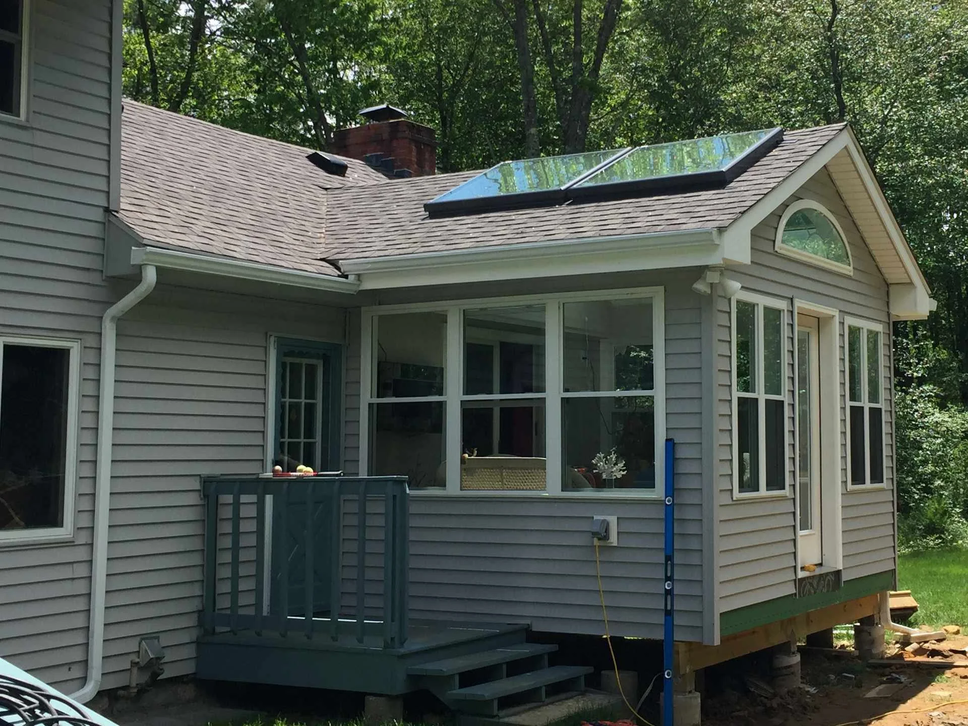 Four-season sunroom addition to house with beige siding, large windows, a small deck with stairs, and skylights on the roof, surrounded by trees.